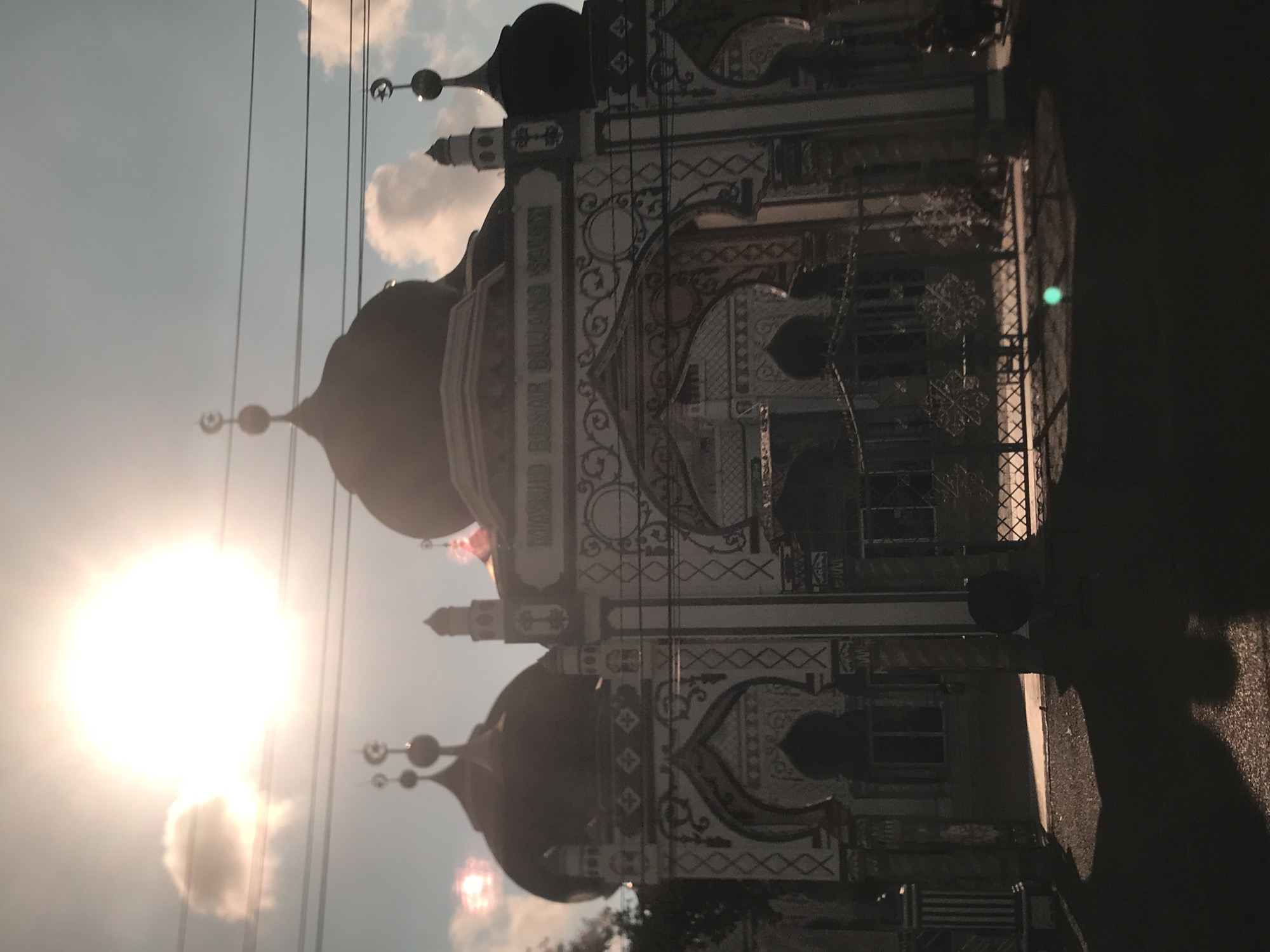 The image shows a mosque in Keudekruenggeukueh, Indonesia, with domes and decorative detailing under a bright sun and cloudy sky. Power lines are visible in the sky. The mosque's architecture features intricate patterns. The sun is positioned to the left, casting a bright glow. Shadows are present on the right side of the image. The image is rotated.