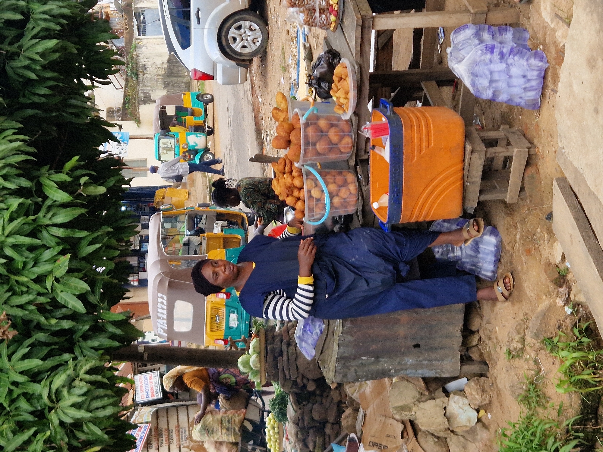 An outdoor market scene in an urban environment features a woman standing behind a stall displaying fried food items and beverages. She is wearing a dark blue overall and a striped shirt with black and white horizontal stripes, and a yellow collar. In front of her, a large orange cooler is placed on a wooden stool. Several transparent buckets filled with fried dough balls are arranged on a wooden table, along with a plate of similar fried items and a black plastic bag. To the right, stacks of plastic-wrapped water bottles are visible on the ground. In the background, a yellow and brown auto-rickshaw is parked, and a silver car is visible. The market appears to be located under a large tree with green foliage. The ground is dusty and scattered with rocks and debris.