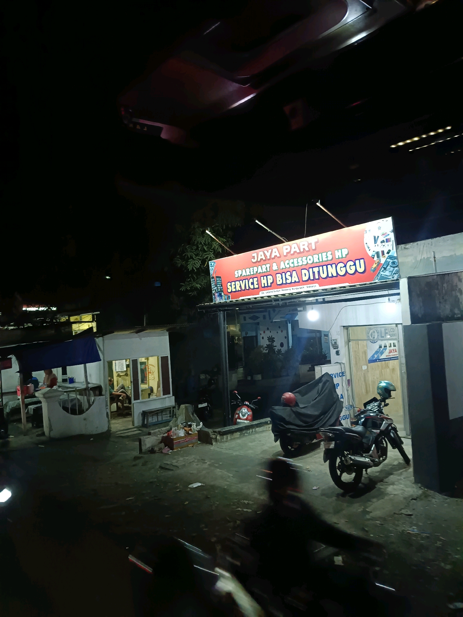 The image shows a night scene outside a shop named "JAYA PART SPAREPART & ACCESSORIES HP SERVICE HP BISA DITUNGGU" located in Bungurasih, Indonesia. The shop is illuminated by a large, bright sign with red and yellow lettering. Below the sign, the shop entrance is visible, and a person is seated inside. To the left of the shop, there's a small food stall with two people present. Several motorcycles are parked in front of the shop, some covered with tarps, and one is being ridden by a blurred figure, indicating movement. The ground is a dark, unpaved surface, and trees are visible in the background. The overall impression is that of a busy, functional commercial area at night.