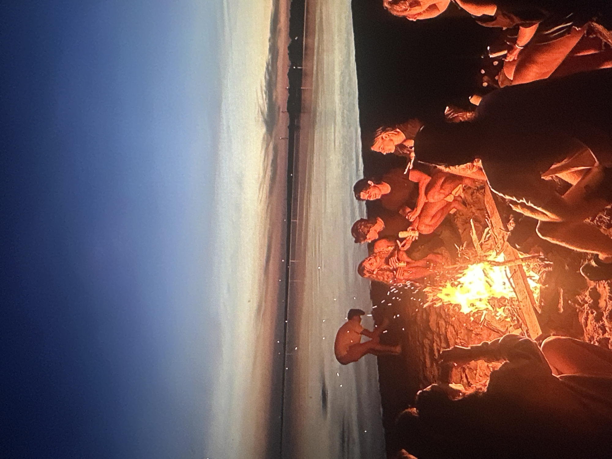 A group of young people are gathered around a bonfire on a sandy beach, situated along the coast of the Baltic Sea. The time appears to be dusk or early evening, with the sky transitioning from a deep blue overhead to softer hues of orange and pink near the horizon. A vast expanse of calm water stretches out before them, reflecting the colors of the sky. In the distance, a faint outline of land and possibly some structures can be discerned.
Several individuals are seated or crouching around the fire, which is actively burning and casting a warm glow on their faces and the surrounding sand. One person, wearing a light-colored shirt, is bent over near the water's edge, seemingly attending to something on the ground. Others are engaged in conversation or simply observing the flames. Some appear to be holding drinks.
The beach itself is composed of fine sand, with scattered pieces of driftwood or branches that likely serve as fuel for the bonfire. The overall atmosphere suggests a casual, social gathering. The location is identified as the beach on the coast of Tallinn, Estonia.