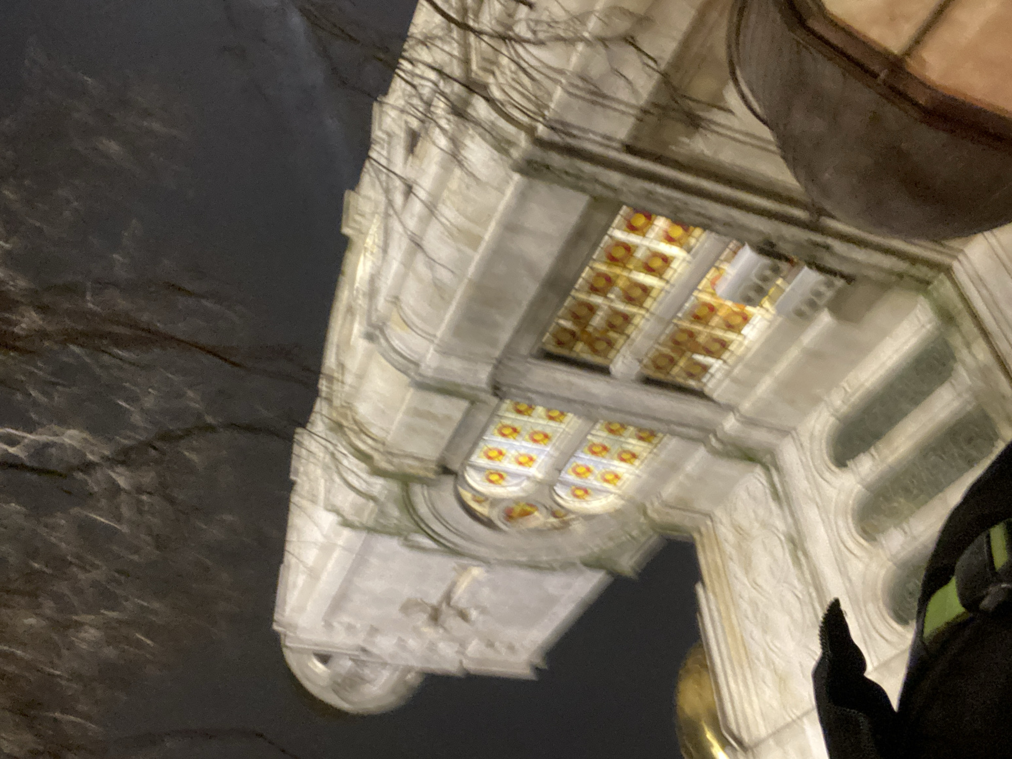 The image shows a low-angle view of a building facade at night. The architecture features ornate stonework, arches, and windows with decorative stained glass. Part of a round, golden dome is visible near the bottom. The building is likely the Dormition of the Theotokos Cathedral in Varna, Bulgaria, known for its elaborate architectural details. A person wearing a black jacket is on the right side of the frame, partially obscuring the view. Bare tree branches are visible on the left side against the night sky.