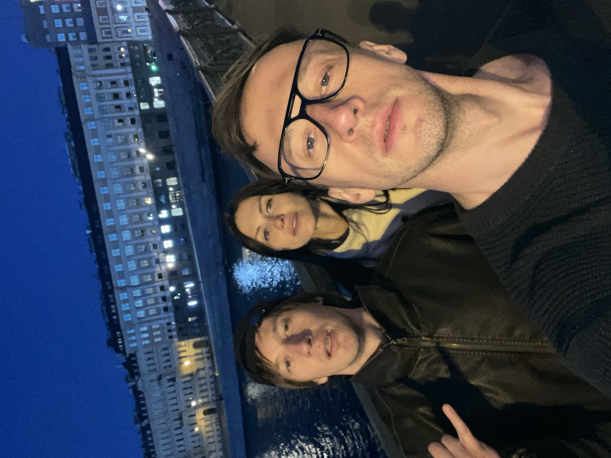 In an outdoor, nighttime selfie, three individuals are pictured with a city skyline in the background. The person in the foreground, on the right, is a man wearing glasses and a dark sweater. To his left and slightly behind him is a woman wearing a light-colored hoodie. On the far left is another man wearing a black jacket and a baseball cap. He is making a hand gesture with his right hand. The background shows a body of water, a bridge railing, and buildings illuminated by lights. The overall setting appears to be a waterfront area in a city at night.