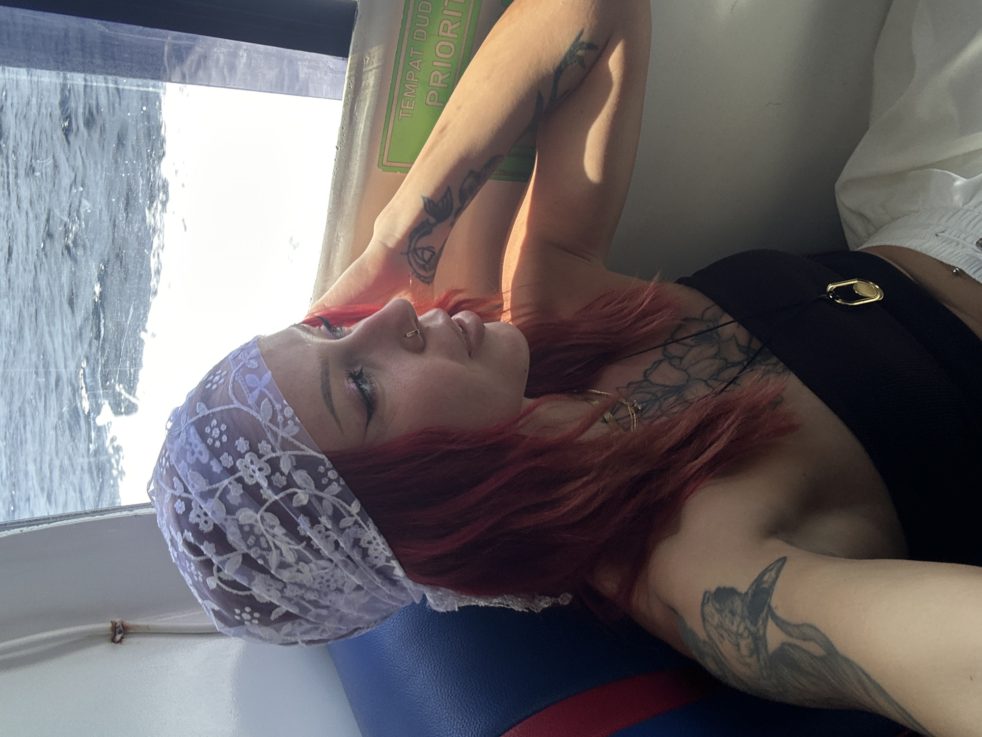 A woman with red hair and tattoos is reclining on a blue and red cushioned seat on what appears to be a boat or ferry. She's wearing a purple lace head covering and a black top with a gold ornament near the collar. A window beside her shows moving water. A green sign with "TEMPAT DUDUK PRIORITI" in white text is also visible next to the window.
The location, Unknown, Indonesia, is derived from the text on the sign, which contains Indonesian words.