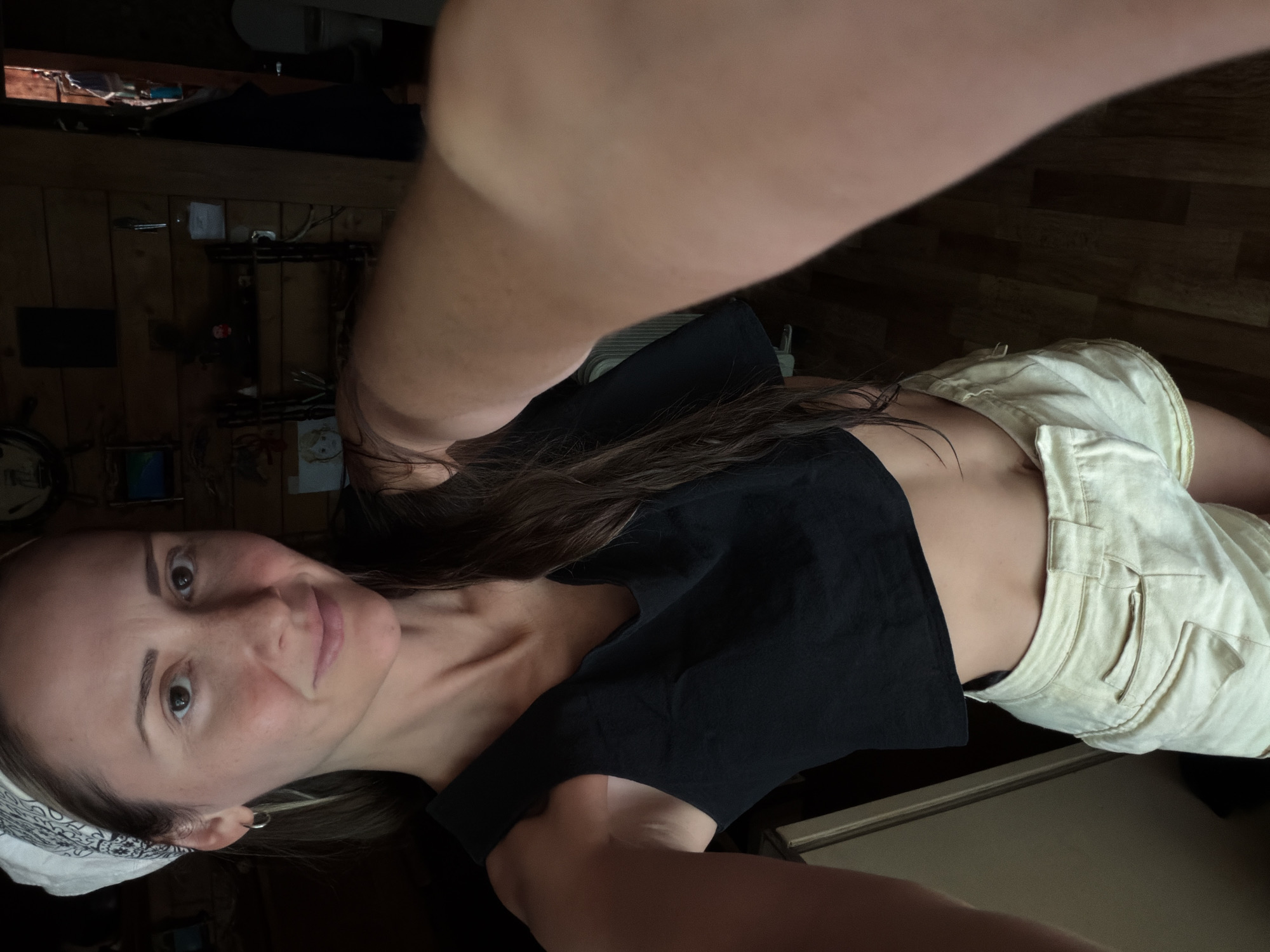 A woman is taking a selfie in a room. She is wearing a black crop top and light-colored shorts. Her hair is long and brown, and she has a bandana on her head. She is smiling and looking at the camera. The room appears to be a living space, with wooden walls and floor. There are shelves on the wall with various items, including what looks like decorative objects and some books. A clock is visible on the left side of the frame. The lighting suggests it might be daytime, or the room is well-lit. The overall mood appears casual and relaxed.