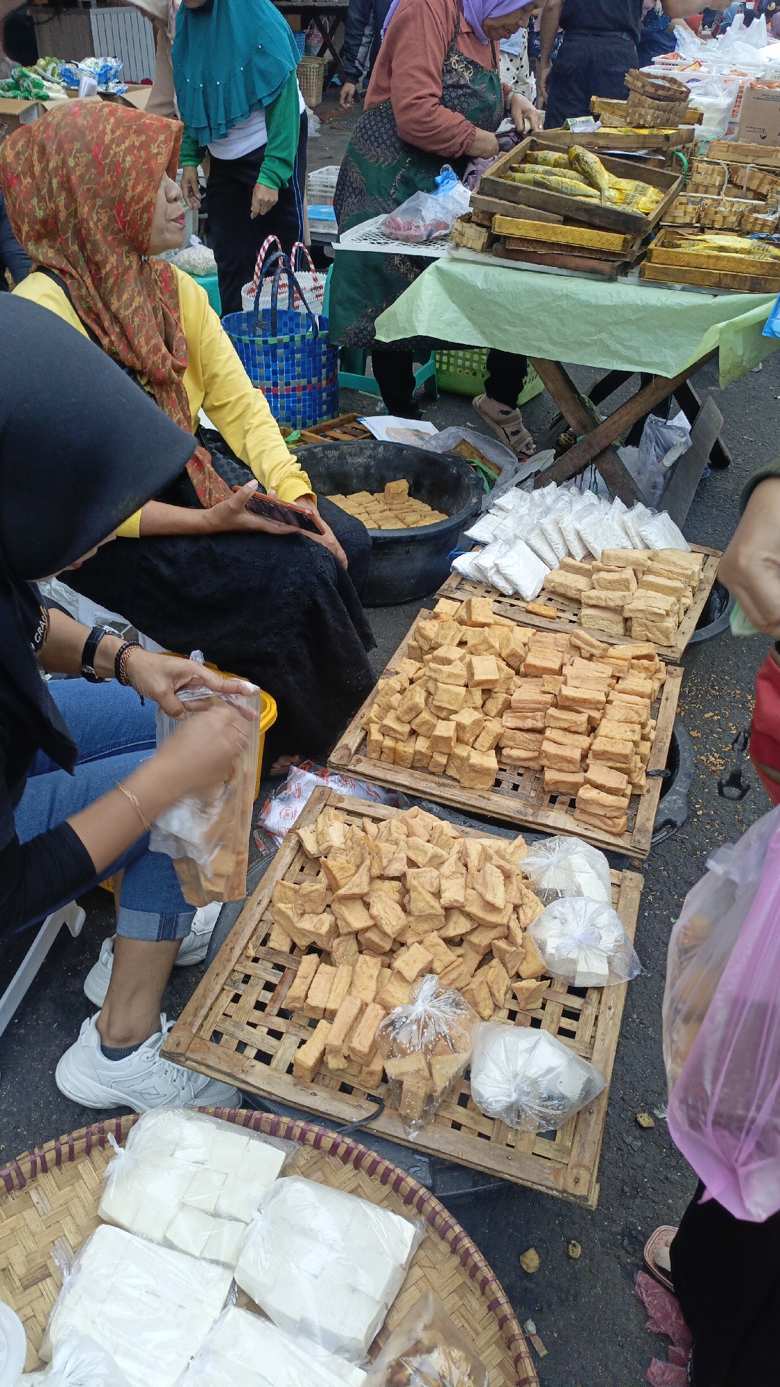 The image depicts a street market scene in Salatiga, Indonesia. Several individuals are present, primarily women, engaged in various activities. In the foreground, a woman wearing blue jeans and a black top is seen packaging cubed, fried tofu into a plastic bag. Beside her, on a woven basket, are packages of white tofu. Behind her, a woman wearing a yellow long-sleeved shirt and a patterned headscarf is seated and looking to the left, holding a smartphone.
In the middle ground, wooden crates are filled with numerous pieces of fried tofu. Another large black basin also contains a substantial amount of fried tofu. To the right, a woman wearing a green apron and a purple headscarf is standing behind a table covered with a green cloth, seemingly attending to customers or arranging goods. The table displays wooden crates filled with what appear to be fish, some of which are yellow.
The background shows more market stalls and other people, suggesting a busy commercial environment. The ground surface is asphalt, and there are various containers and packaging materials scattered around. The overall impression is of a typical Indonesian food market.
