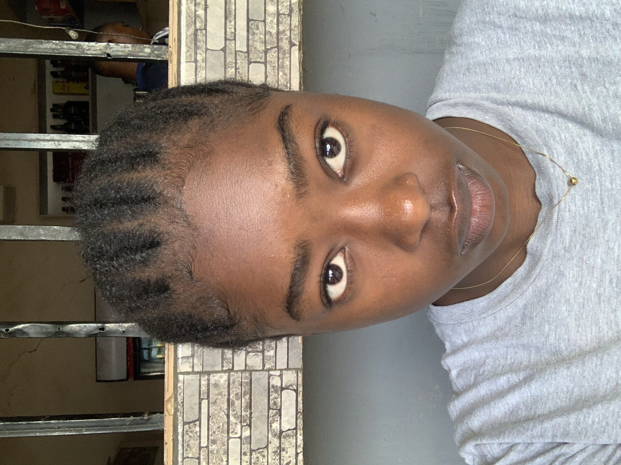 A medium shot of a person with dark skin, brown eyes, and tightly braided dark hair, angled slightly to the right. The person is wearing a light gray T-shirt and a gold necklace. The background is a mix of a gray wall or surface and a textured brick-like wall. Through a metal grate in the background, a glimpse of an interior space with shelves and various objects can be seen. The location cannot be determined with only this image.