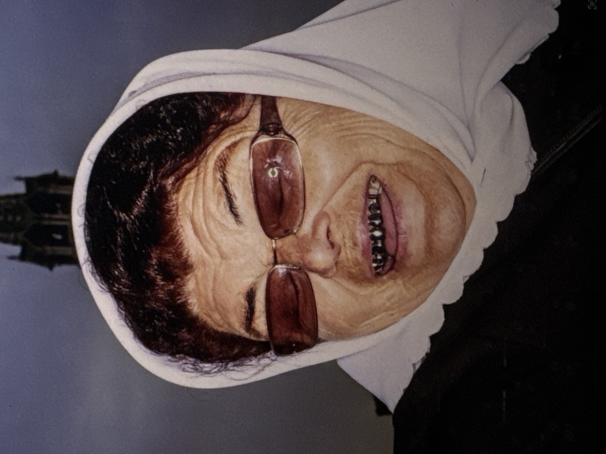 A close-up, medium shot photograph captures a woman wearing a white headscarf and dark sunglasses. She is looking directly at the camera with her mouth open, revealing gold-capped teeth. The woman's face shows wrinkles, and her hair is dark and curly. Behind her, a blurred, dark, gothic-style building with spires is visible against a dark, overcast sky. The overall lighting is dim, suggesting an outdoor setting under cloudy conditions. There are no visible indications of the specific location being Kuta Selatan, Indonesia.