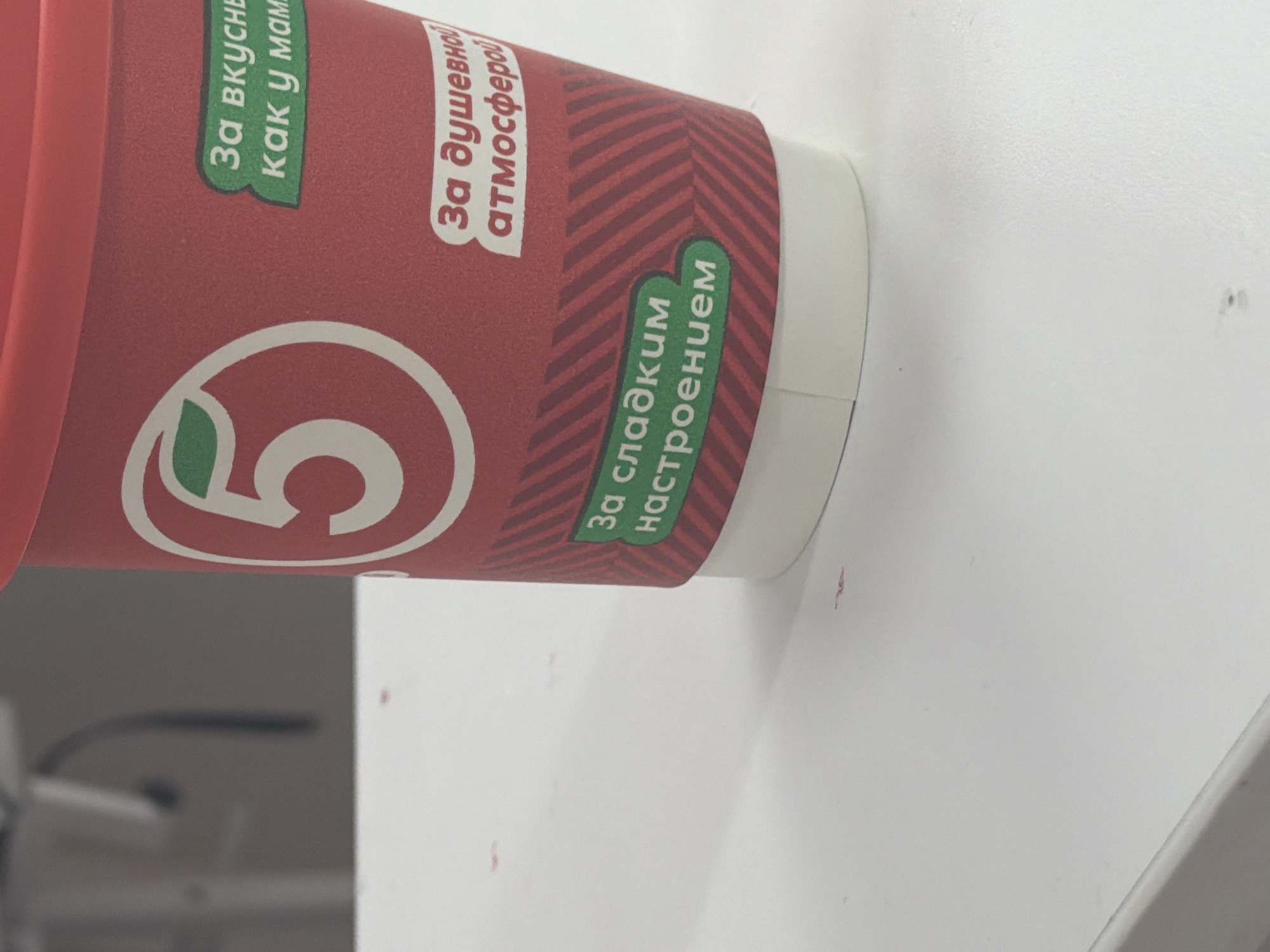 A close-up, eye-level shot shows the side of a red disposable coffee cup with a white rim and bottom, resting on a white table. The cup features a large white number "5" inside a white circle with a green leaf at the top left. Text in green is visible on the cup. Above the number "5," a green sticker reads "За вкусны как у мам" which translates to "For tasty like mom's". Below the number "5," another green sticker reads "За душевной атмосферой" which translates to "For a soulful atmosphere". Further down the cup, a green banner reads "За сладким настроением" which translates to "For a sweet mood". The background is blurred and appears to be an indoor setting with neutral colors.