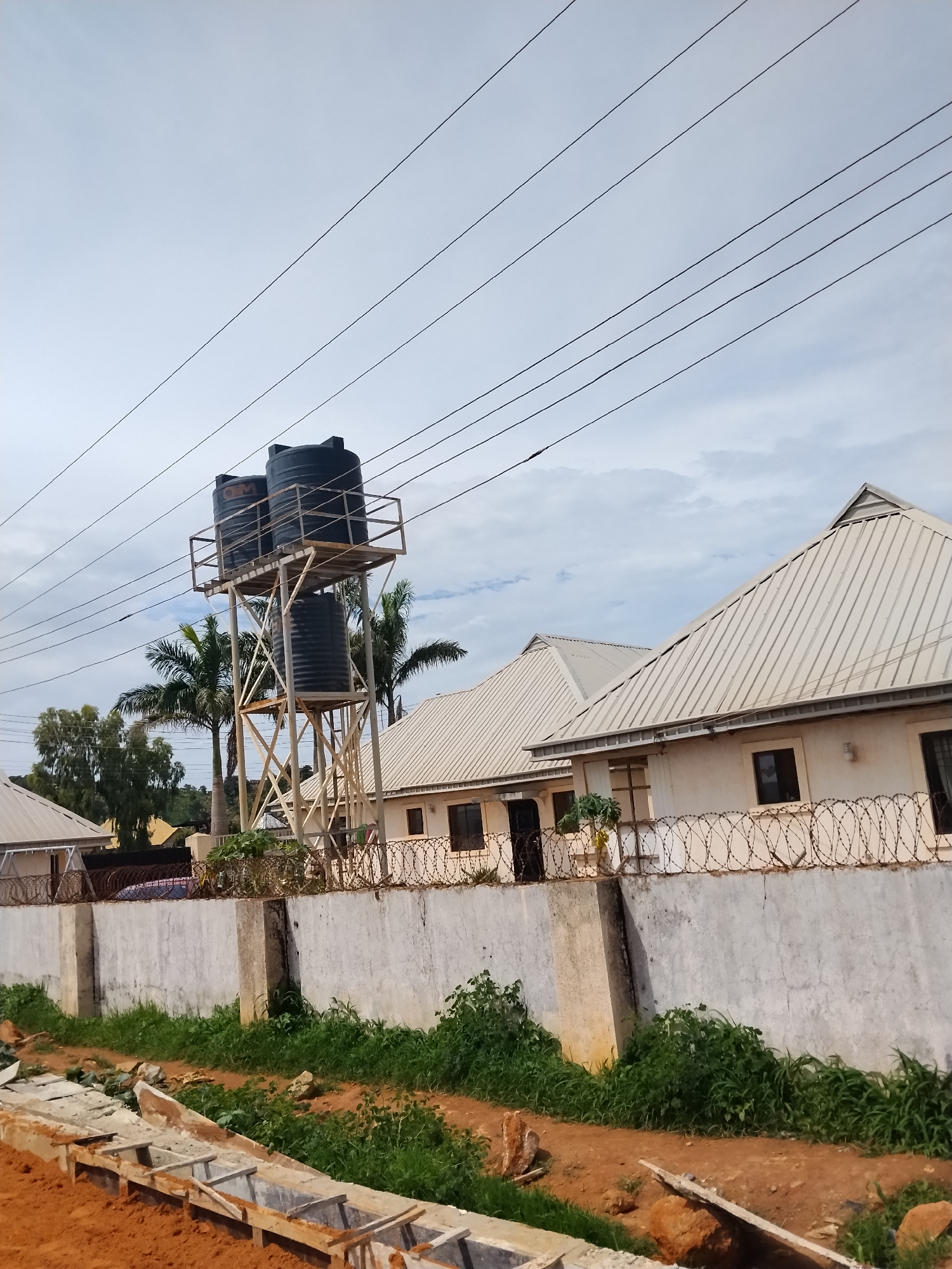 The image depicts a residential area in Laminga, Nigeria, characterized by a cluster of single-story buildings with corrugated metal roofs. Prominently featured is a tall metal water tower supporting three large black water tanks, situated behind a concrete wall topped with barbed wire. Power lines crisscross the sky above. In the foreground, construction appears to be underway with exposed concrete structures and red earth. Lush green vegetation lines the area between the construction site and the wall, with palm trees visible behind the water tower. The sky is overcast with clouds. No people are visible in the scene.