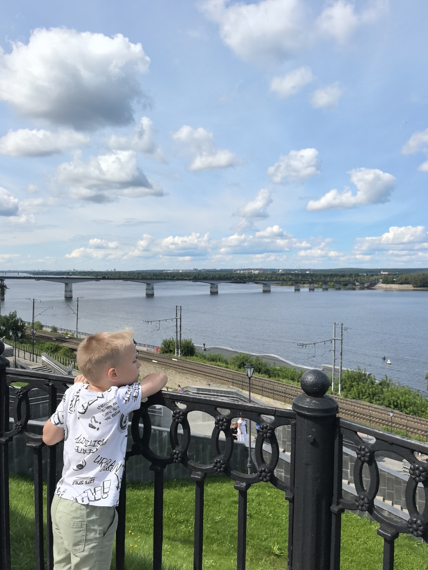 In Perm, Russia, a young boy with blond hair leans on a black metal railing, gazing out at a wide river under a partly cloudy sky. The railing features decorative circular and floral patterns. Behind the boy, a set of train tracks runs parallel to the riverbank, bordered by lush green grass and trees. In the distance, a long bridge with multiple piers spans the river, connecting the two sides of the city. Power lines and poles are visible along the riverfront. The scene captures a moment of quiet observation and contemplation by the river.