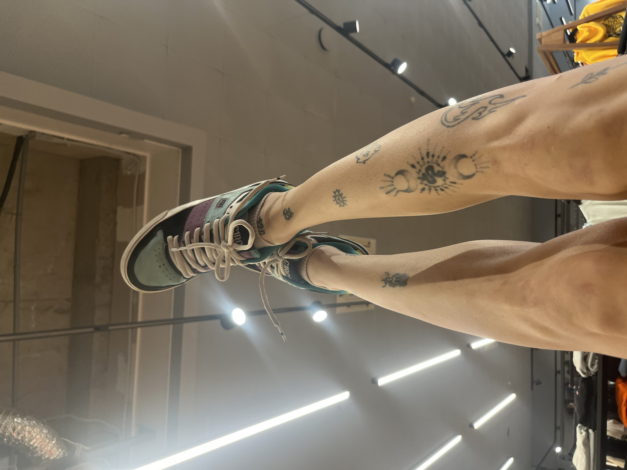 The image shows two legs with multiple tattoos wearing sneakers. The left sneaker is green, purple, and white. The background consists of a store with ceiling lights and a wall with a doorframe. Clothing racks are visible on the right side. The image was likely taken in a store in the city of Krasnoyarsk, Russia.