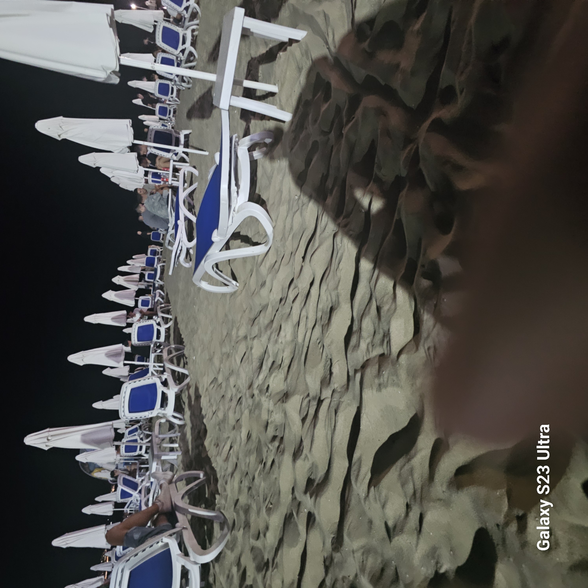 The image depicts a nighttime scene on a sandy beach, likely in Nesebar, Bulgaria, given the provided watermark. Numerous white beach umbrellas are set up, some open and others closed, casting shadows on the sand. White plastic sun loungers with blue cushions are arranged in rows. In the background, several people are visible sitting on loungers or at small tables. One person's legs are prominently featured in the foreground, resting on a sun lounger. The sand shows clear patterns of ripples and shadows, indicating it is a beach environment. The overall atmosphere is calm and the lighting suggests artificial illumination from nearby establishments.