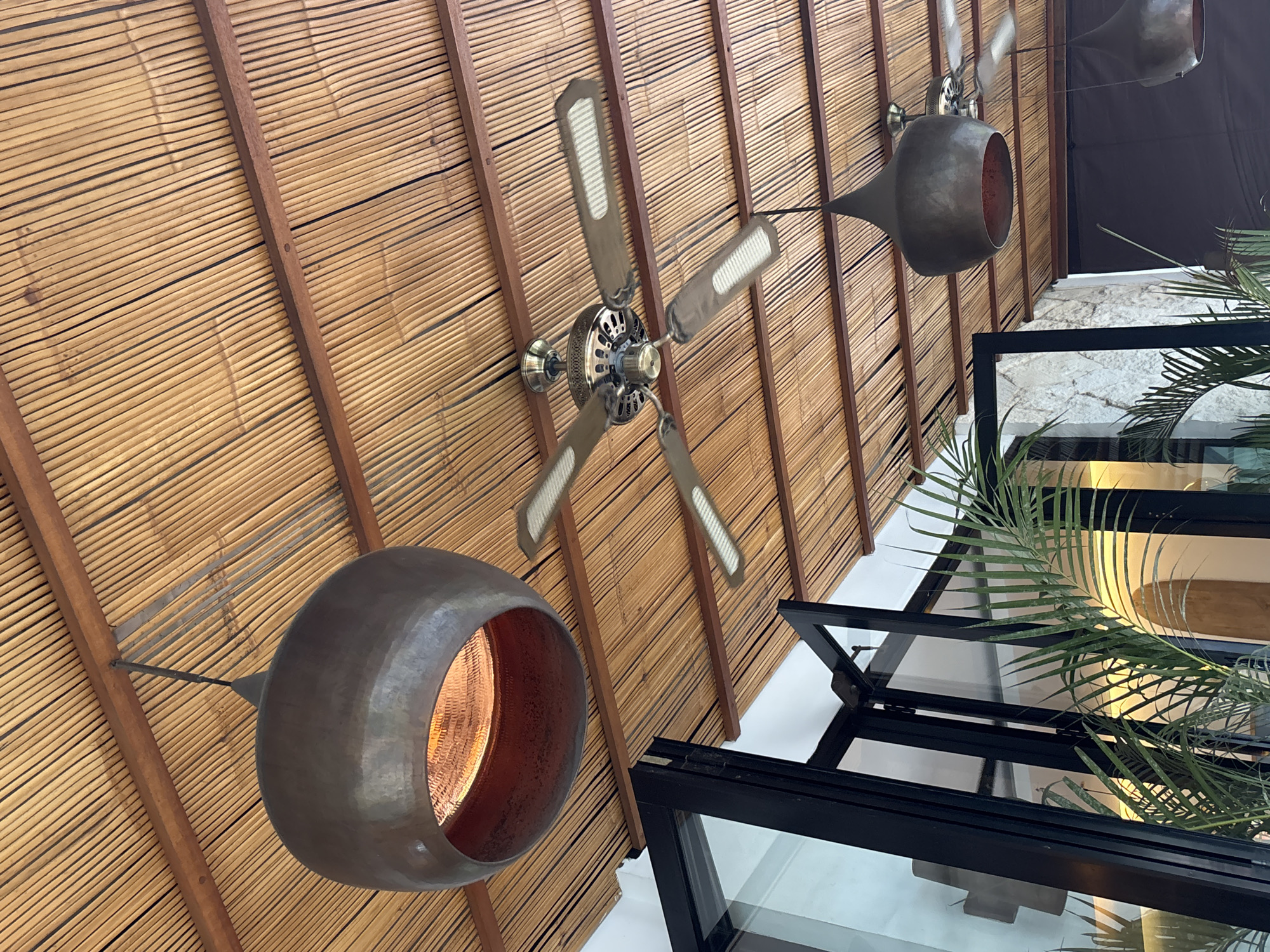 The image shows an indoor space in Kuta Selatan, Indonesia, featuring a ceiling made of bamboo and dark wood supports. Two ceiling fans with five blades each are visible. There are two hanging light fixtures with large, round, copper-colored shades. The lower right of the image shows a black-framed window with multiple panes and green palm fronds. The background appears to be a building with white walls.