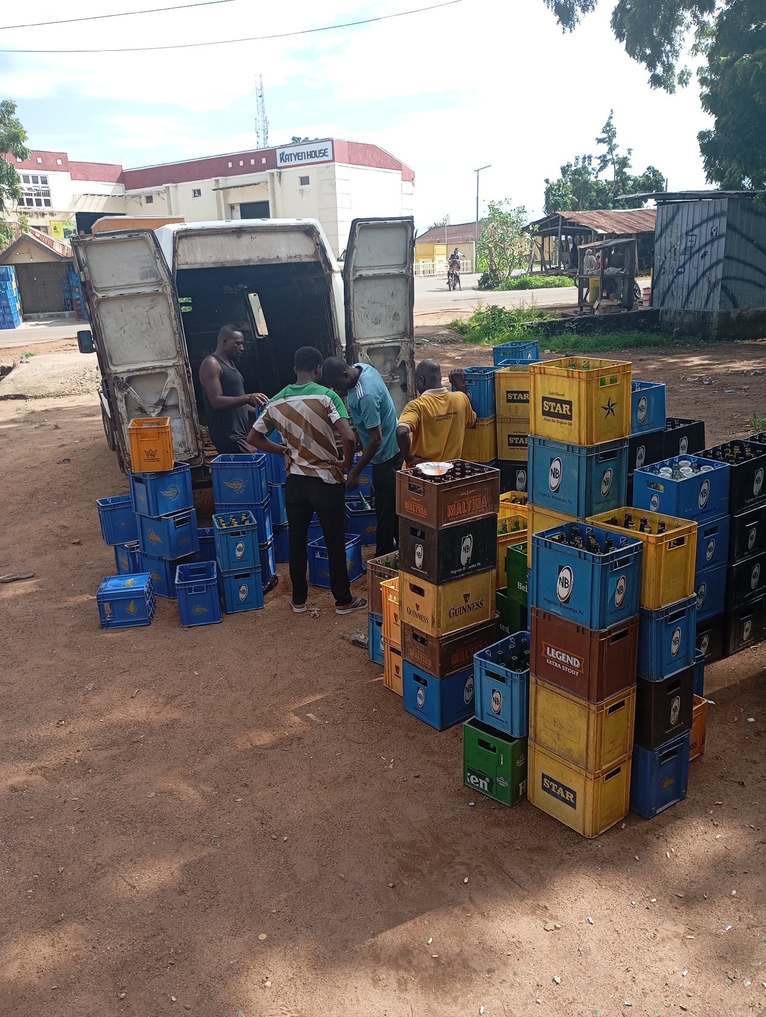 In Shendam, Nigeria, at an outdoor location with a dirt ground, a white van with its rear doors open is positioned. Several individuals, appearing to be workers, are engaged in unloading and arranging numerous crates of beverages. The crates are predominantly blue, yellow, and brown, with some displaying labels such as "STAR," "MALTINA," "GUINNESS," "LEGEND," and "NB Nigerian Breweries Plc." Some crates are stacked precariously high, containing bottles. In the background, a building with the signage "KATYEN HOUSE" is visible, along with other structures and trees. The sky is partly cloudy.
