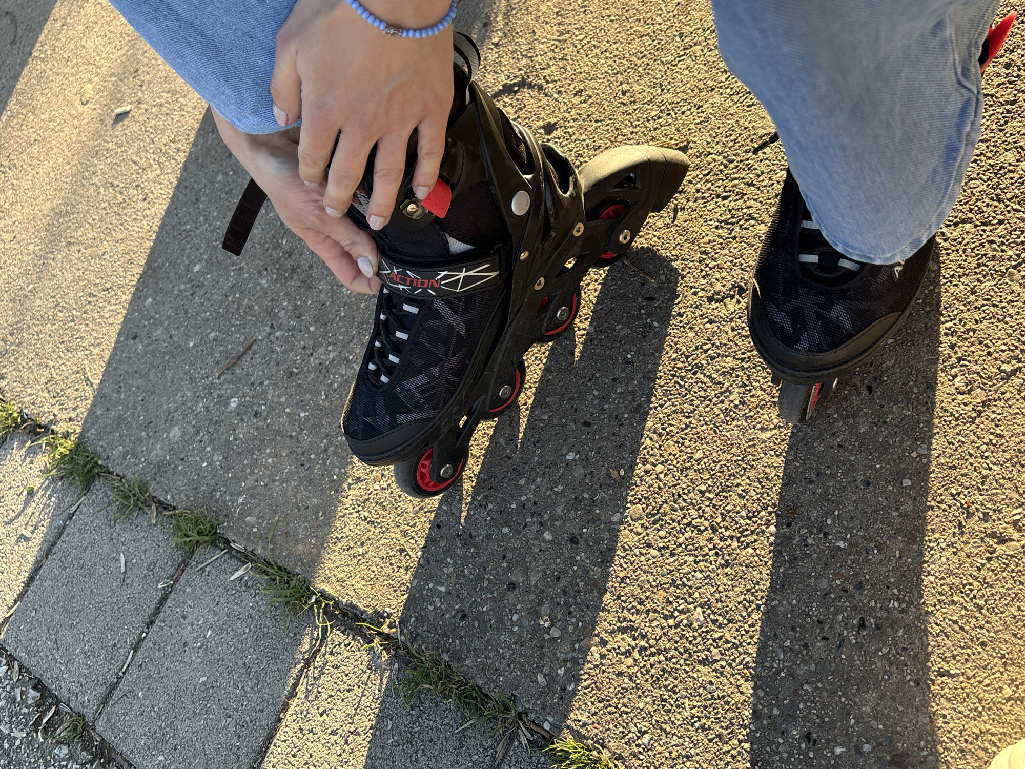 A person is shown putting on inline skates outside on a sunny day. The person is wearing light blue jeans and a beaded bracelet. The inline skates are black with red wheels. One skate is on and the person is fastening the closure of the other skate. The location is outdoors on a concrete surface with grass growing in between the cracks. I have no information about the location in the city of Kragujevac, Serbia.
