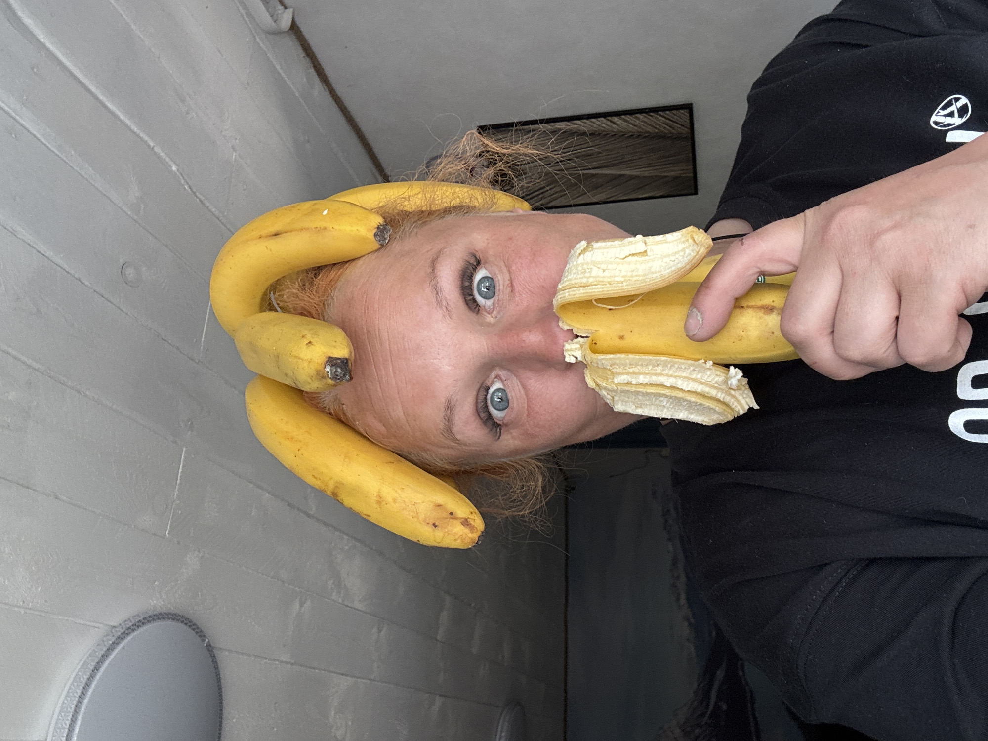 A fair-skinned person with blue eyes and reddish-blonde hair is holding a partially peeled banana to their mouth and has three other bananas on their head. They are wearing a black t-shirt with a white design. The background consists of a white wall with horizontal panels and a dark rectangular object. The location is not identifiable from the image.