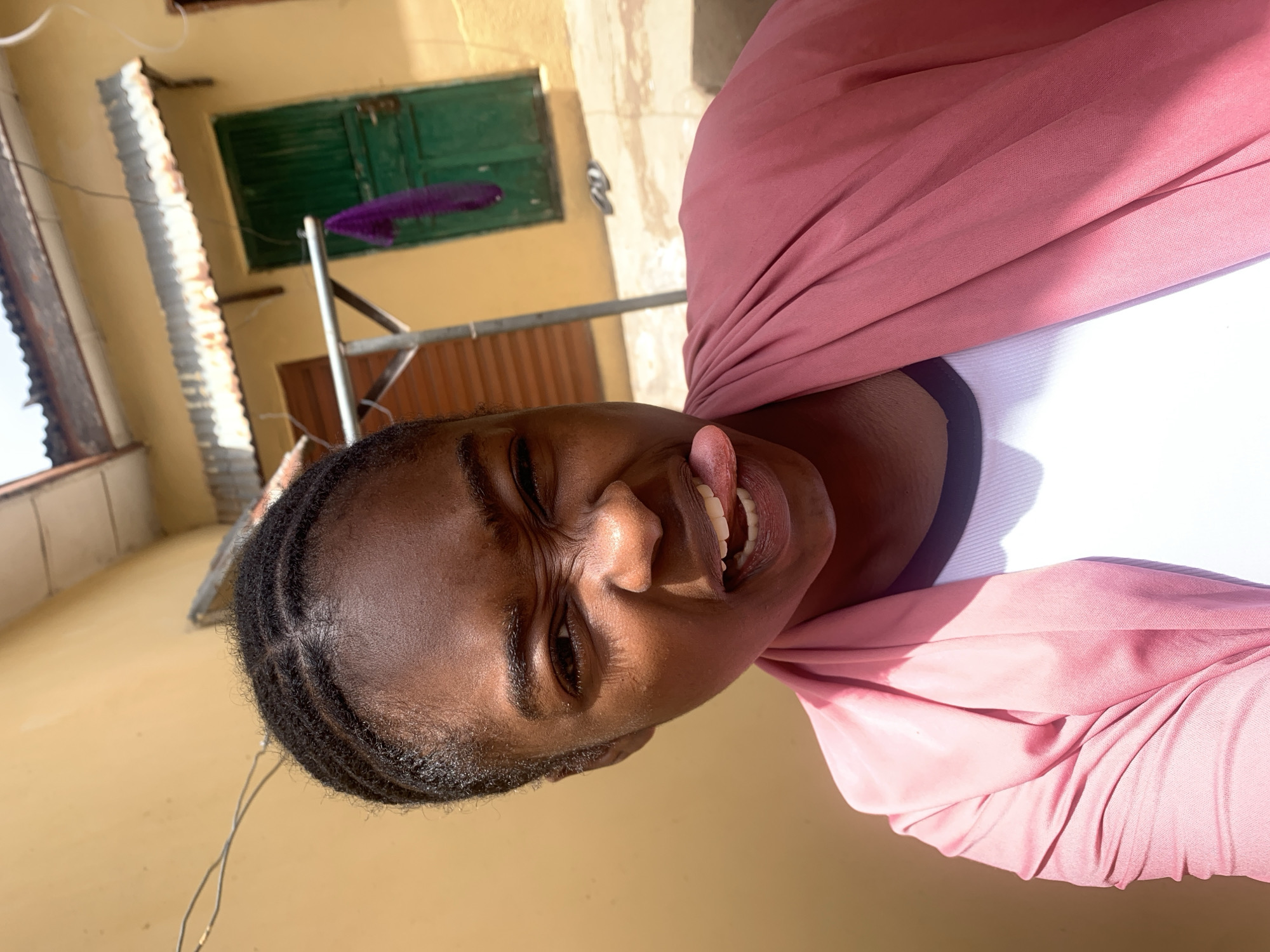 A young African woman poses outdoors in Rafin Par, Nigeria. She is wearing a white top with a black trim, covered by a pink draped fabric. Her hair is styled in braids close to her scalp. She is sticking out her tongue and squinting. In the background, a building with green shutters and a metal clothesline are visible. The surroundings suggest a residential area in Rafin Par, Nigeria.