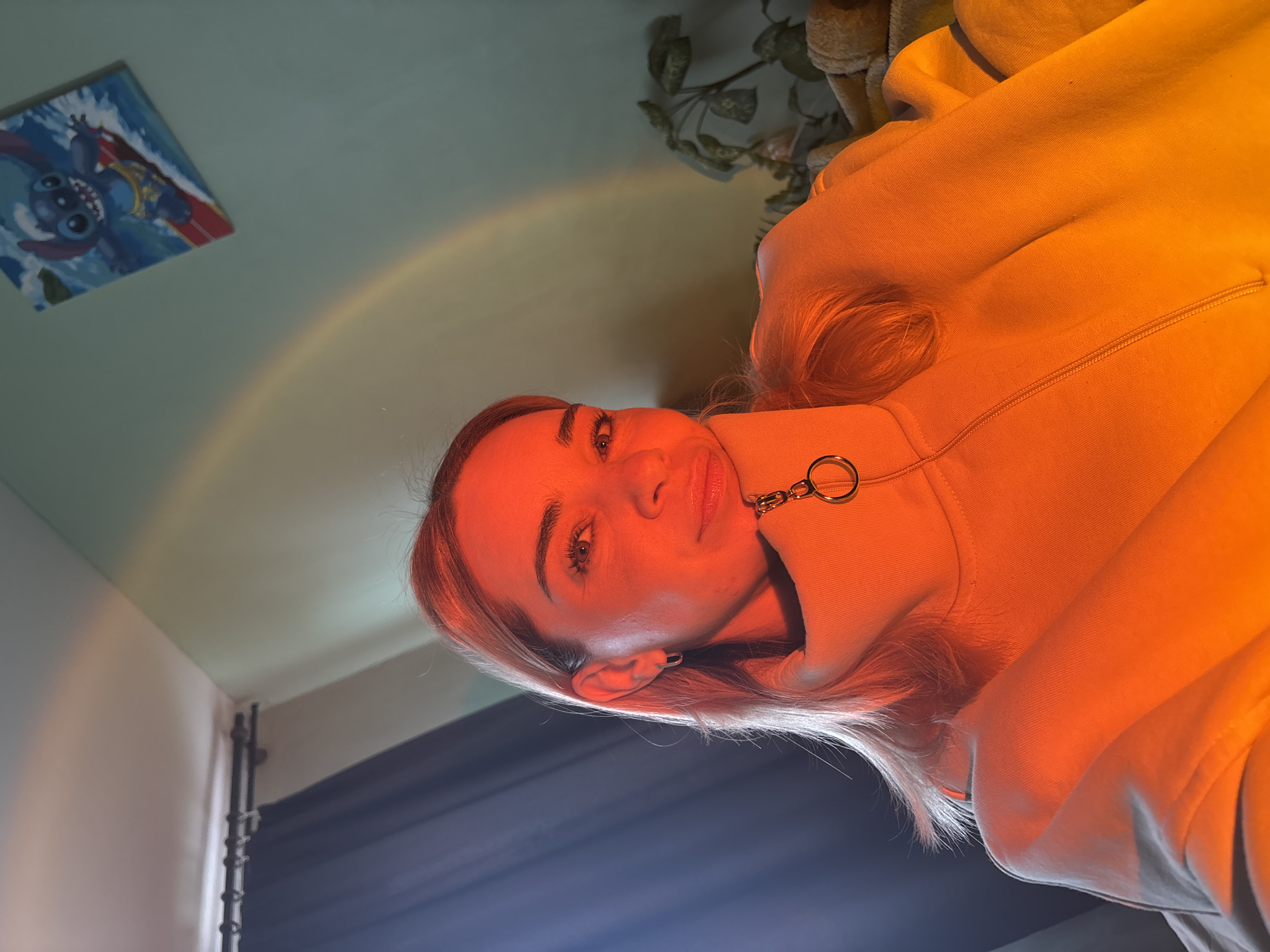 A portrait-style photograph, rotated 90 degrees counter-clockwise, presents a fair-skinned woman wearing a light-colored, zip-up hooded sweatshirt with a circular zipper pull. She is looking directly at the camera. The lighting has an orange hue, casting shadows and highlights across her face and clothing.
In the background, above and behind the woman, the ceiling and upper wall are visible. A faint rainbow-like arc is cast on the ceiling. To the left, a square painting or print featuring the character "Stitch" from the Disney film "Lilo & Stitch" hangs on the wall. In the upper right corner of the frame, a plant sits on top of what appears to be an upholstered object. The lower left corner of the image reveals a dark vertical stripe, possibly a window frame or edge of a wall. The setting appears to be indoors.