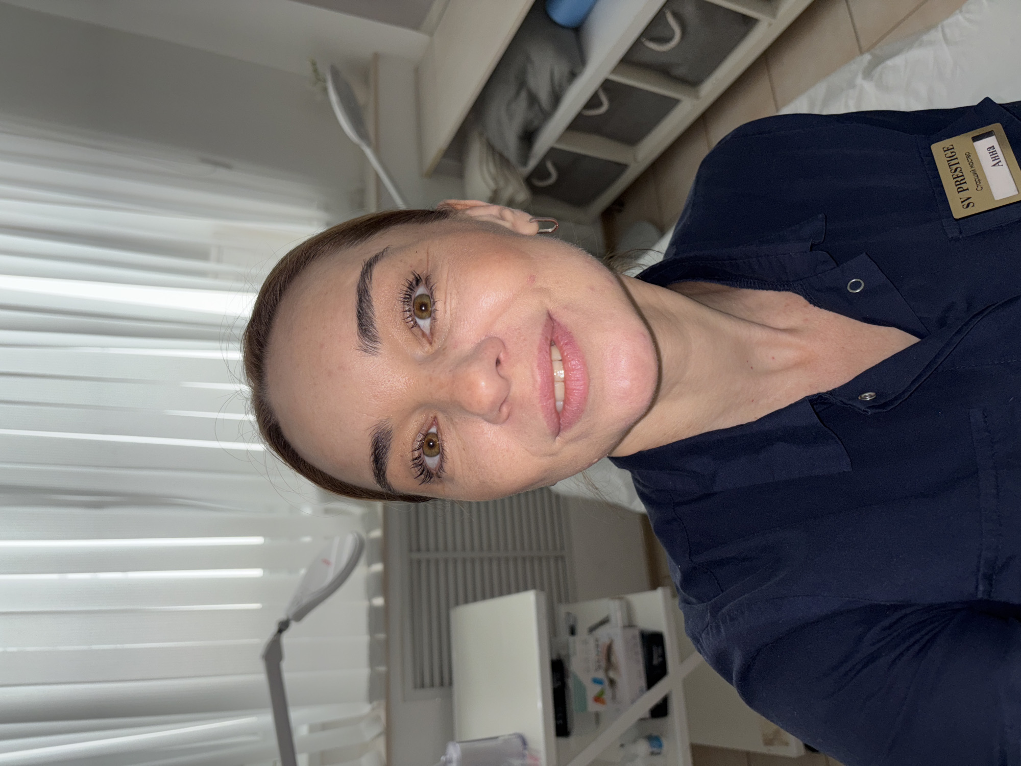 A woman with brown hair and eyes smiles towards the camera. She wears a dark blue collared shirt with a name tag that reads "SV PRESTIGE" and "AHHA." The environment appears to be a clinical or office space, indicated by the presence of blinds, a medical lamp, and shelving with various items. It is unknown if the woman is in Vsevolozhsk, Russia.