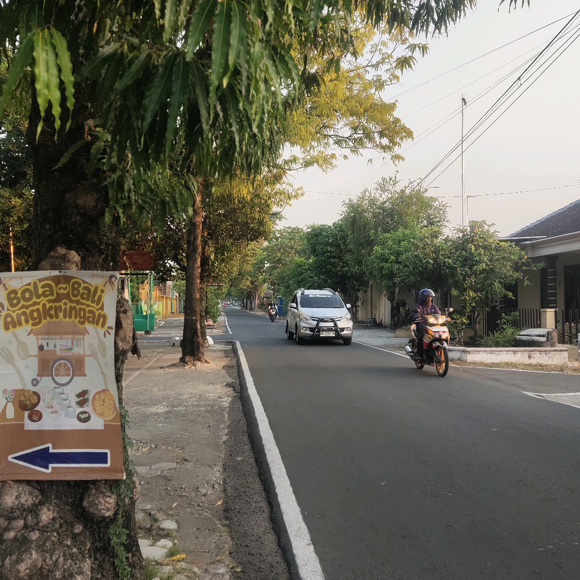The image depicts a street scene in Sragen, Indonesia, likely in the early morning or late afternoon based on the lighting. A prominent sign attached to a tree on the left side advertises "Bola~Bali Angkringan," featuring illustrations of food items and a food cart, with a blue arrow pointing left. The street is paved, with a white line marking the edge. A silver car is traveling down the road, and a person on a motorcycle is also visible. Lush green trees line both sides of the street, creating a shaded pathway. Houses with tiled roofs and walls are visible on the right side of the road. The overall atmosphere suggests a typical residential or commercial street in a warm climate.