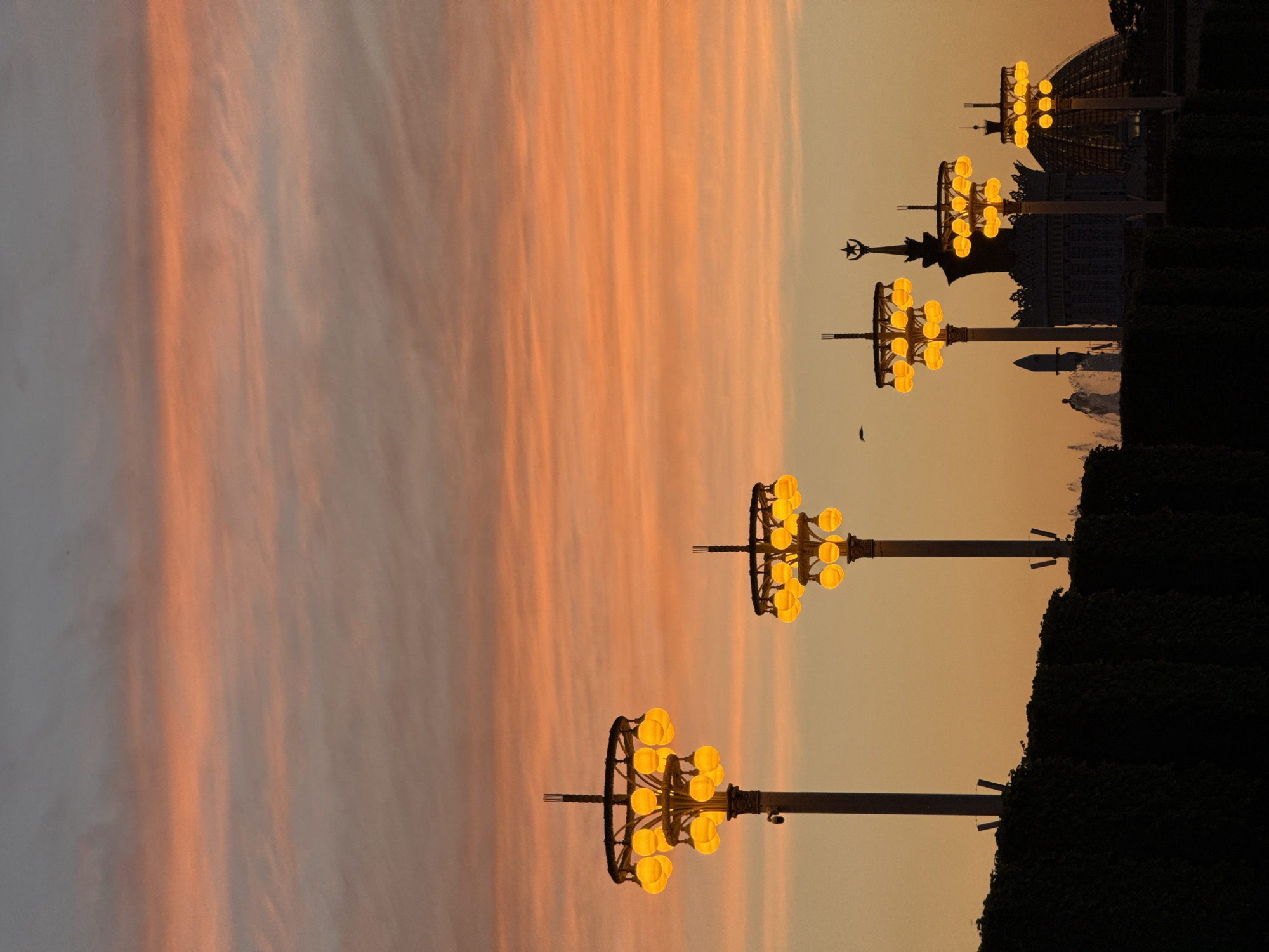 The image captures a serene evening scene at Exhibition of Achievements of National Economy (VDNKh) in Moscow, Russia. The sky displays a gradient of colors, from orange to light yellow. In the foreground are ornate, vintage-style lampposts with multiple globe-shaped lights. These are spaced out along the edge of dark green bushes, creating a strong contrast between the illuminated lamps and the shadowed foliage. The building in the background is the Cosmos Pavilion, or Space Pavilion. There are no people visible, and the focus is primarily on the architecture and the natural light. A dark silhouette of a bird is visible in the center.