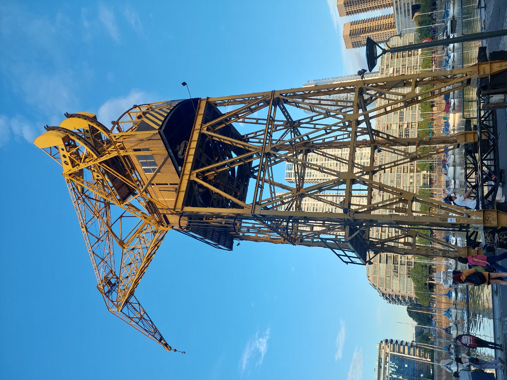 A tall, yellow crane stands prominently against a blue sky in Puerto Madero, Buenos Aires, Argentina. The crane is composed of a lattice-like steel framework, featuring a cabin near the top and a long, extending arm. Tall buildings are visible in the background, alongside a waterfront lined with sailboats and other watercraft. People are seen walking along the edge of the water, some interacting with the environment. The scene appears to be captured on a sunny day.