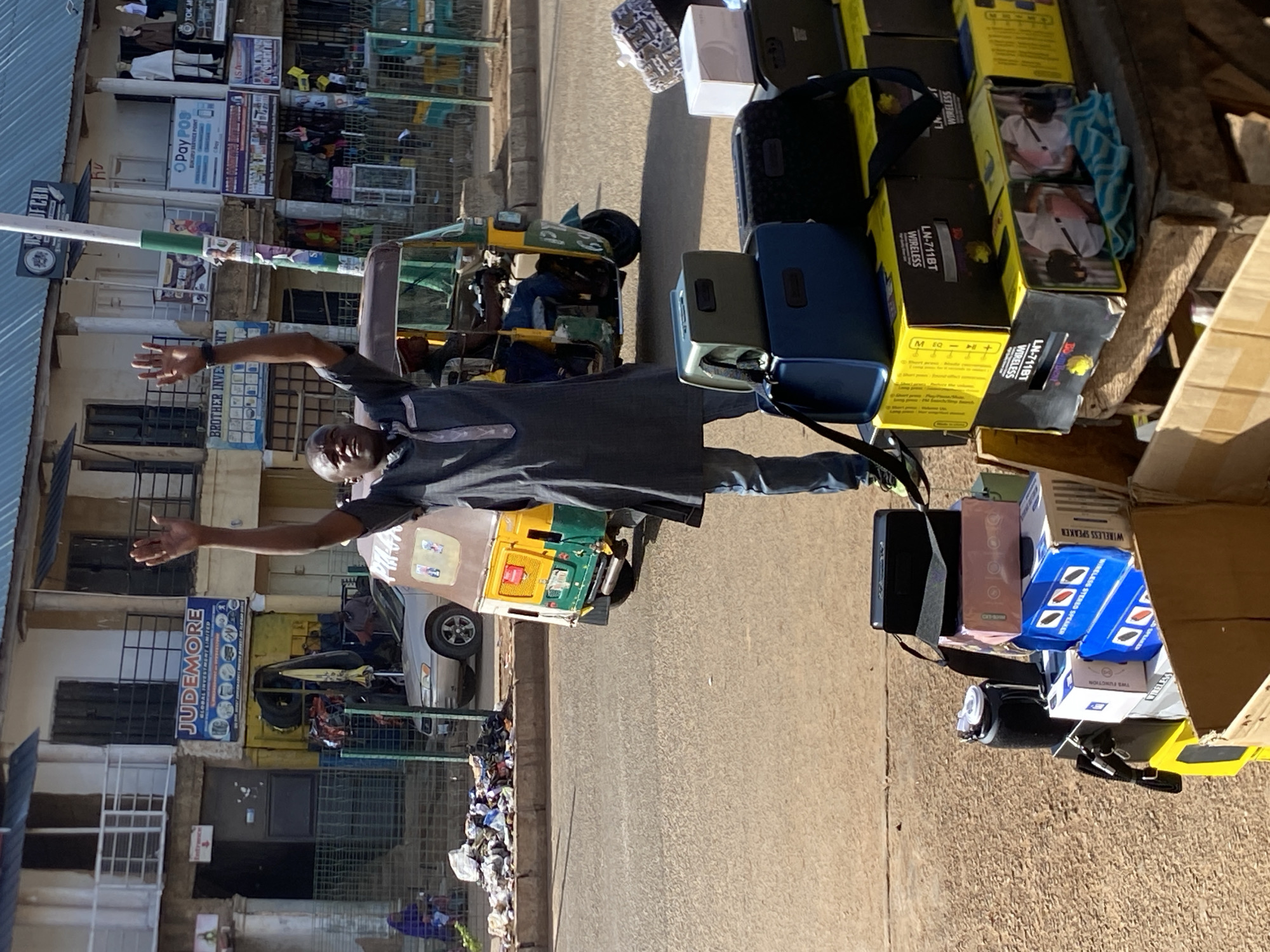 An image captures a lively street scene in Bukuru, Nigeria, likely a commercial or market area, under bright daylight. In the foreground, a man wearing a dark-colored traditional-style top and dark pants stands with his arms outstretched, smiling warmly towards the camera.
To his right, a street stall is laden with various goods, predominantly electronics. Notable items include numerous yellow boxes labeled "LN-711BT WIRELESS," likely containing headphones or speakers, with images of people wearing them. Other boxes are visible, some mentioning "WIRELESS STEREO SPEAKER" and "TWS FUNCTION." Dark bags and a black speaker are also part of the merchandise.
Behind the man and further down the street, buildings line the road. Several businesses are identifiable by their signs, including "Pay POS Bukuru Service Point," "ISA," "TOK M," "JUDEMORE GLOBAL INVESTMENT LIMITED," and "BROTHER INVES." A green and yellow motorized rickshaw, commonly known as a Keke Napep in Nigeria, is parked near the buildings, and a silver car is also visible. Along the curb, piles of trash or discarded items are evident. The bright sun casts clear shadows, indicating a sunny day. The overall scene depicts a busy, open-air commercial environment.