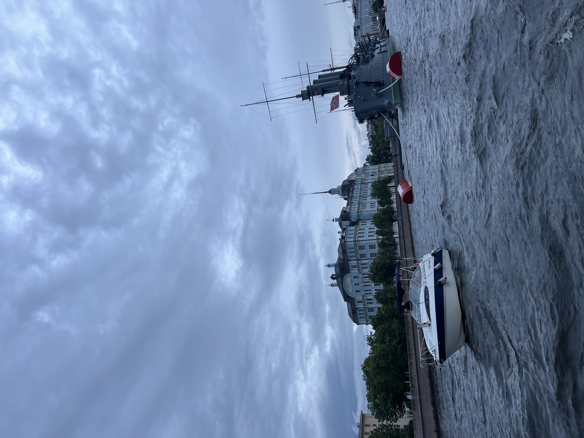 The image shows a wide river in Saint Petersburg, Russia. On the right side of the river, a large, dark gray cruiser warship is moored. The ship has several masts, smokestacks, and a Russian flag flying from its stern. It is tethered by large chains to two large, red and white buoys.
On the left bank of the river, a light blue, ornate building with a dark roof and multiple windows stands behind a line of green trees. Further to the left, a lighter colored building is visible.
In the foreground, a white motorboat with a blue stripe is moving across the river. The boat has a dark canopy and at least two people are visible inside. The sky is overcast with a layer of dark, textured clouds. The river's surface is choppy with small waves.