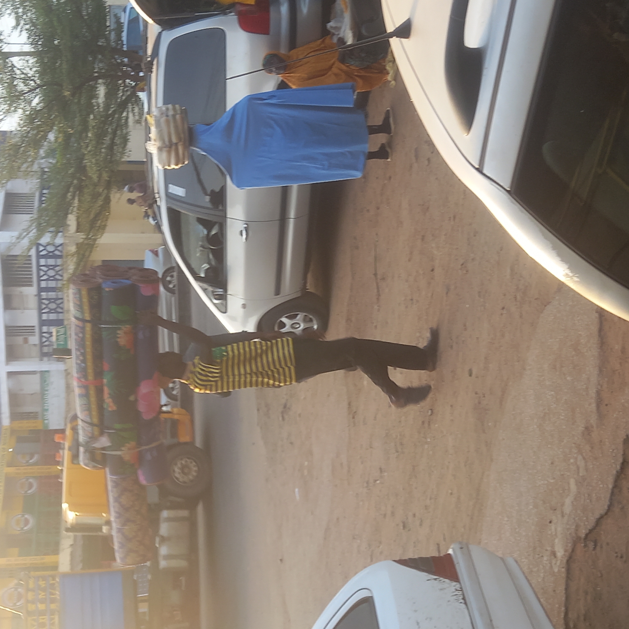 The image depicts a street scene in Dadi Riba, Nigeria. A man wearing a striped shirt and dark pants is carrying a stack of colorful mats on his head. Nearby, a person in a blue garment is carrying a bundle of sticks or similar items. Vehicles, including a white car and a yellow tricycle loaded with more mats, are visible on the street. The setting appears to be a residential area, with buildings and a tree lining the street. The ground is sandy.