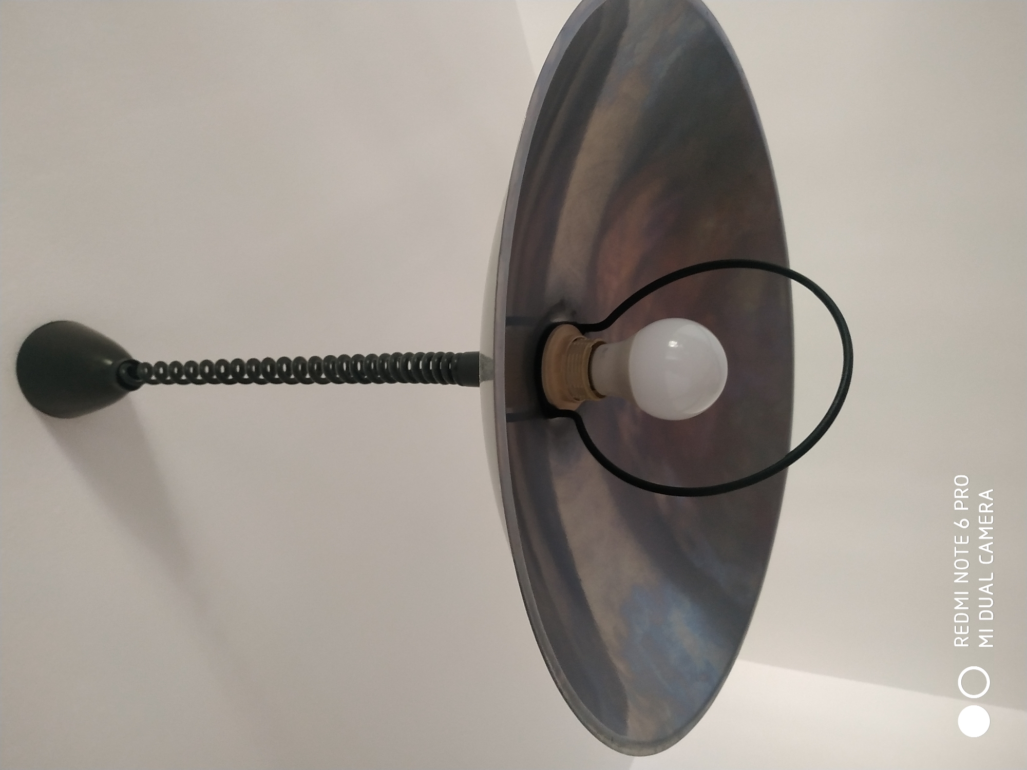 The image shows a ceiling light fixture indoors. The fixture has a black spiral cord connecting a black ceiling mount to a large, metallic, dish-shaped lampshade. A single white lightbulb is visible hanging from a socket within the lampshade. The lampshade has a brushed metal finish with some reflective, swirling patterns. The ceiling is painted white. There is a watermark in the bottom left corner of the image that reads "REDMI NOTE 6 PRO MI DUAL CAMERA". There are no people or animals present in the image. The image does not provide any information about the location in the city of Fryazino, Russia.