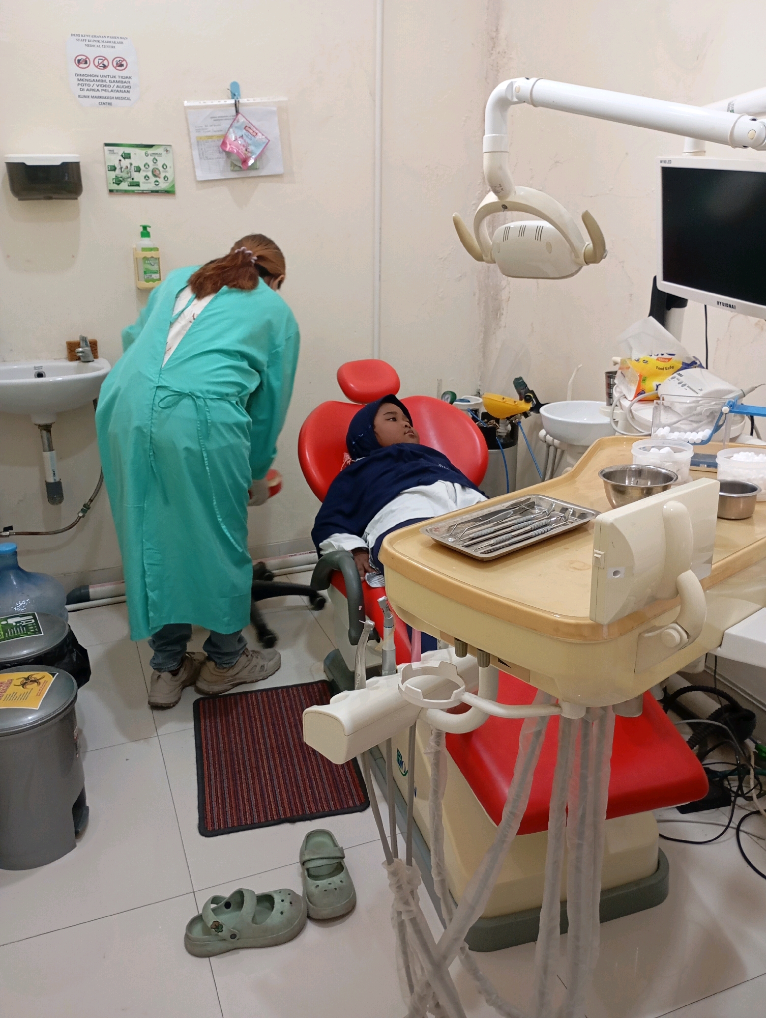 The image depicts a dental clinic in Babelan, Indonesia, likely part of the Mitra Keluarga Medical Centre, as indicated by signage on the wall. A dental professional, wearing a teal gown and gloves, stands near a sink, facing away from the camera. To the right, a young patient, wearing a dark hooded jacket, lies in a red dental chair. A dental examination light hangs above the chair. Various dental instruments are laid out on a tray on a dental unit. The environment appears sterile, with medical supplies and equipment visible. A water dispenser, waste bin, and signage regarding photography restrictions are also present in the room.