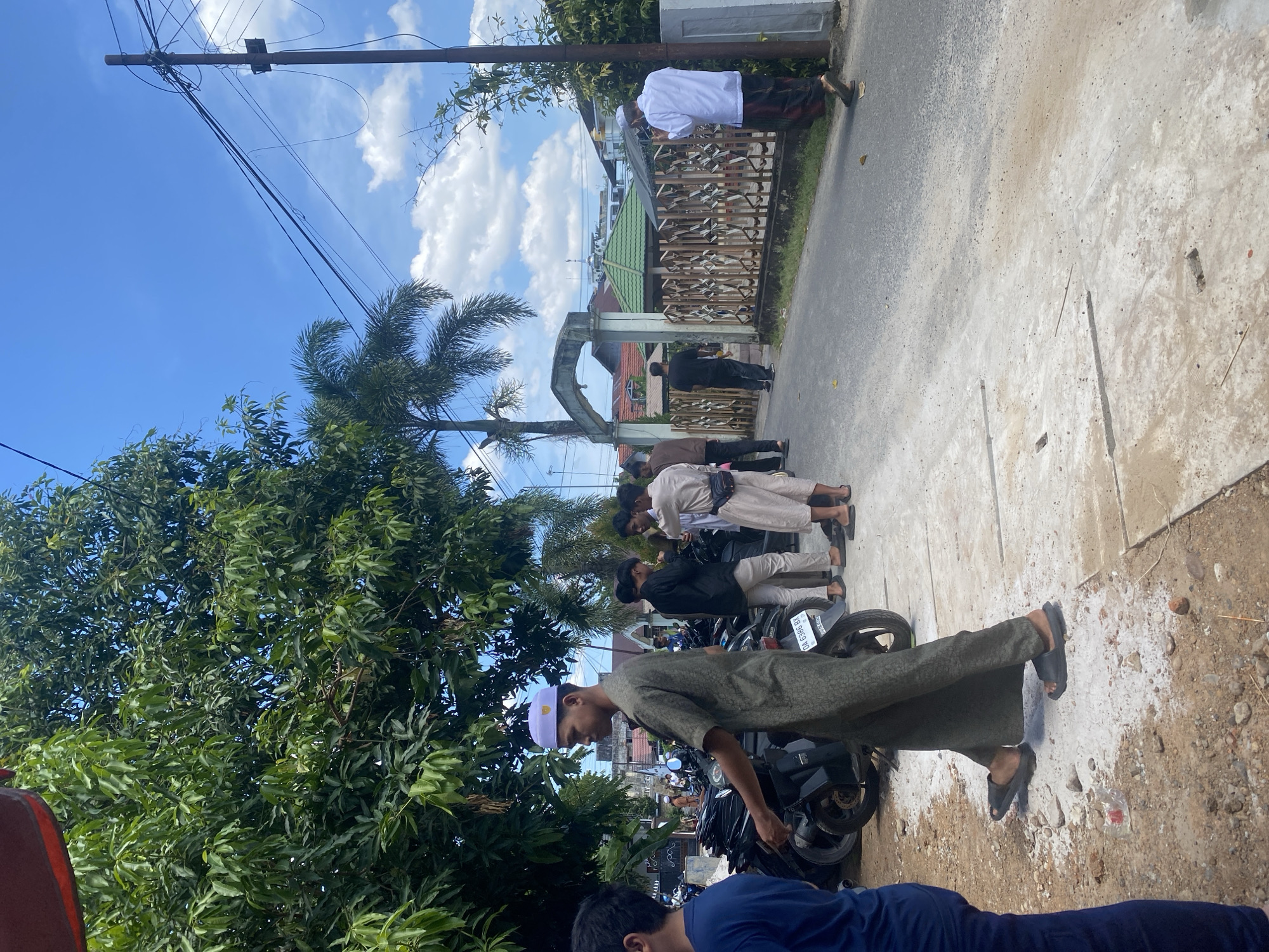 A group of men are gathered on a street in Banjarbaru, Indonesia, under a partly cloudy sky. In the foreground, a man in a green tunic and white cap walks to the left. Several motorcycles are parked on the left side of the street. Further down the street, other men in traditional Islamic attire stand and converse. A decorative gate with a green-roofed structure behind it is visible on the right side of the street, with a man in a white shirt and patterned sarong standing near it. The environment appears to be a residential or community area with trees and buildings in the background.