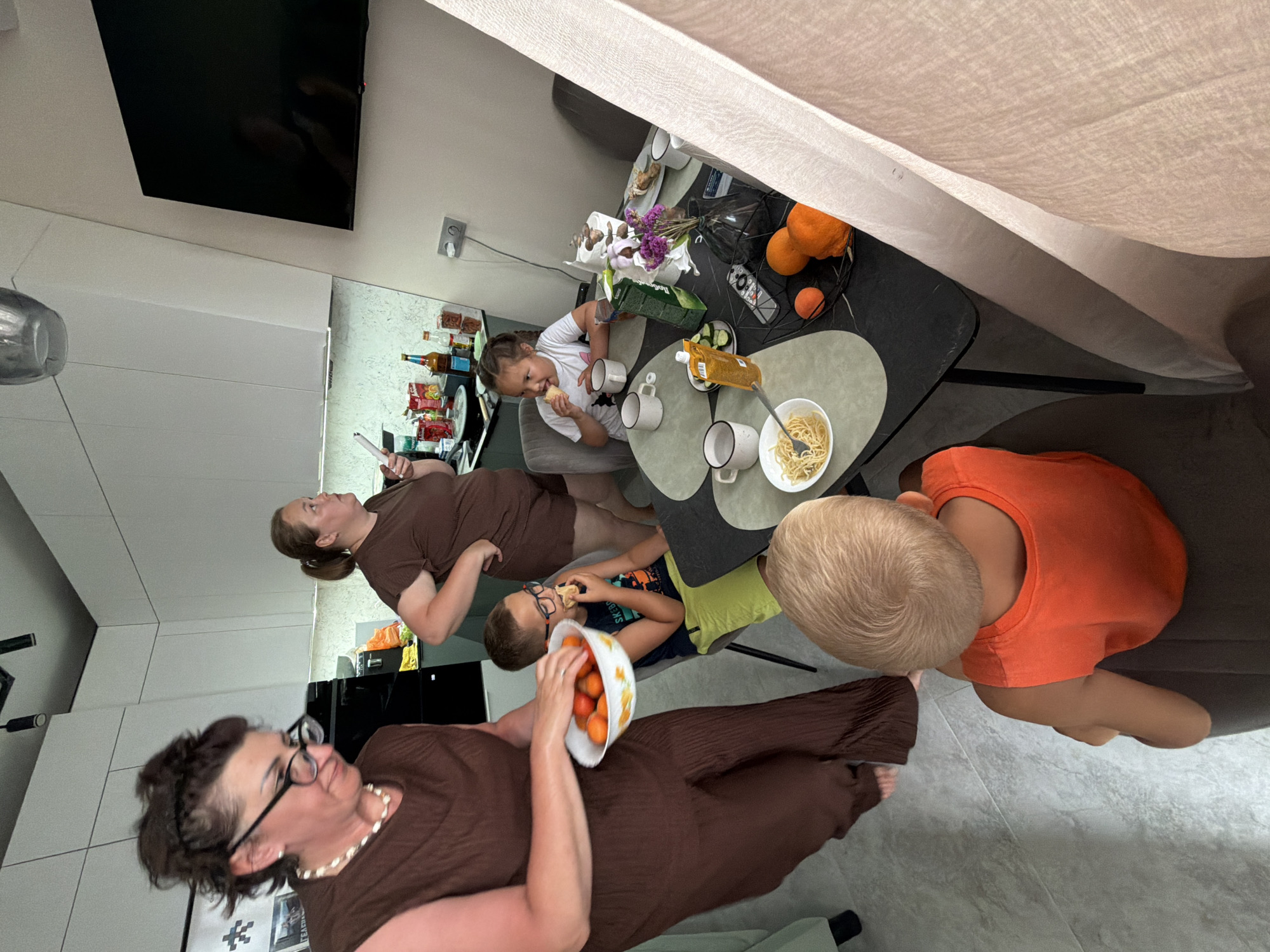 In a modern kitchen, a family is gathered around a dining table. A woman in a brown dress and glasses holds a bowl of oranges, standing near a counter with various food items and appliances. To her right, another woman in a brown top observes the scene. At the table, a boy in glasses with a blue and orange shirt is eating. In front of him, a girl in a white top is eating an ice cream cone. A young boy in an orange shirt is seated in the foreground, facing away from the camera. The table is set with plates of food, bowls of pasta, cups, and a vase of flowers. The kitchen features white cabinets, a black countertop, and a television mounted on the wall. The floor is tiled in a light gray. The overall impression is a casual family meal in a well-lit home environment. The image does not contain any specific visual cues to pinpoint its location within the city of Novorossiysk, Russia.