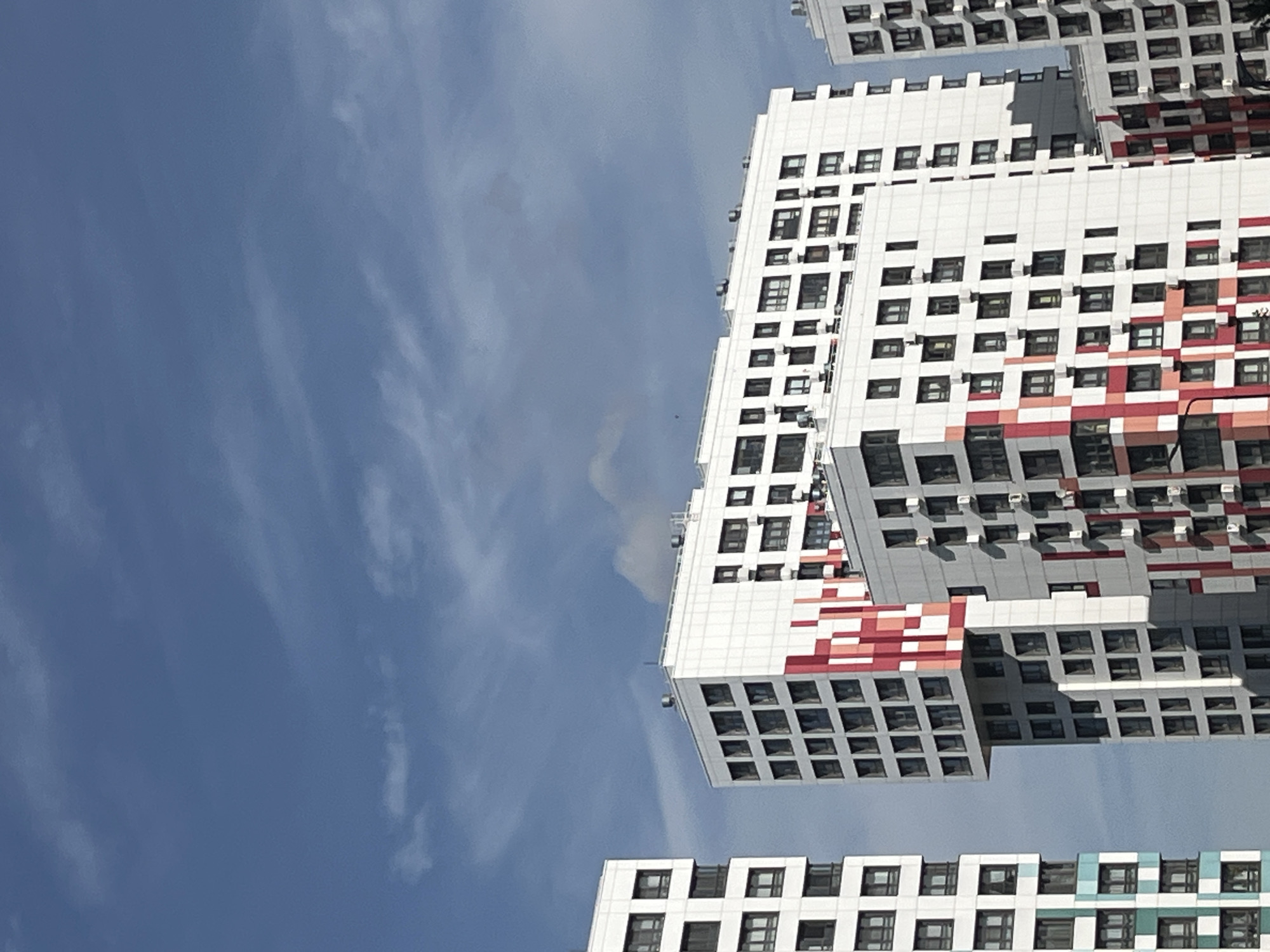 The image shows a cluster of modern residential buildings against a bright blue sky with scattered white clouds. The buildings are tall and appear to be constructed from white panels, with numerous dark-framed windows arranged in a grid pattern. Some of the buildings feature colorful architectural accents, specifically red and white geometric patterns on their facades. The buildings are positioned close together, suggesting a dense urban environment. The perspective is from a low angle, looking upwards at the structures. The scene is devoid of any visible people or vehicles, creating a sense of stillness. The lighting suggests it is daytime, likely with the sun at an angle that casts minimal shadows on the buildings. The overall impression is one of contemporary city living. Based on the architectural style and visual characteristics of the buildings, this scene is likely located in the city of Moscow, Russia.