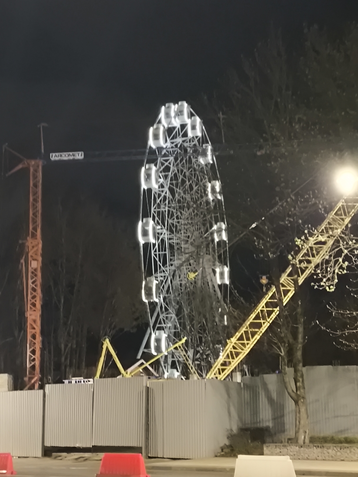 Observed at night, the image reveals a Ferris wheel illuminated with lights in Smolensk, Russia. Two cranes flank the Ferris wheel, one painted orange and the other yellow. A gray metal fence runs along the lower part of the frame, and a tree stands to the right of the yellow crane. The environment includes bare trees and a street or open area with red and white traffic barriers in the foreground.