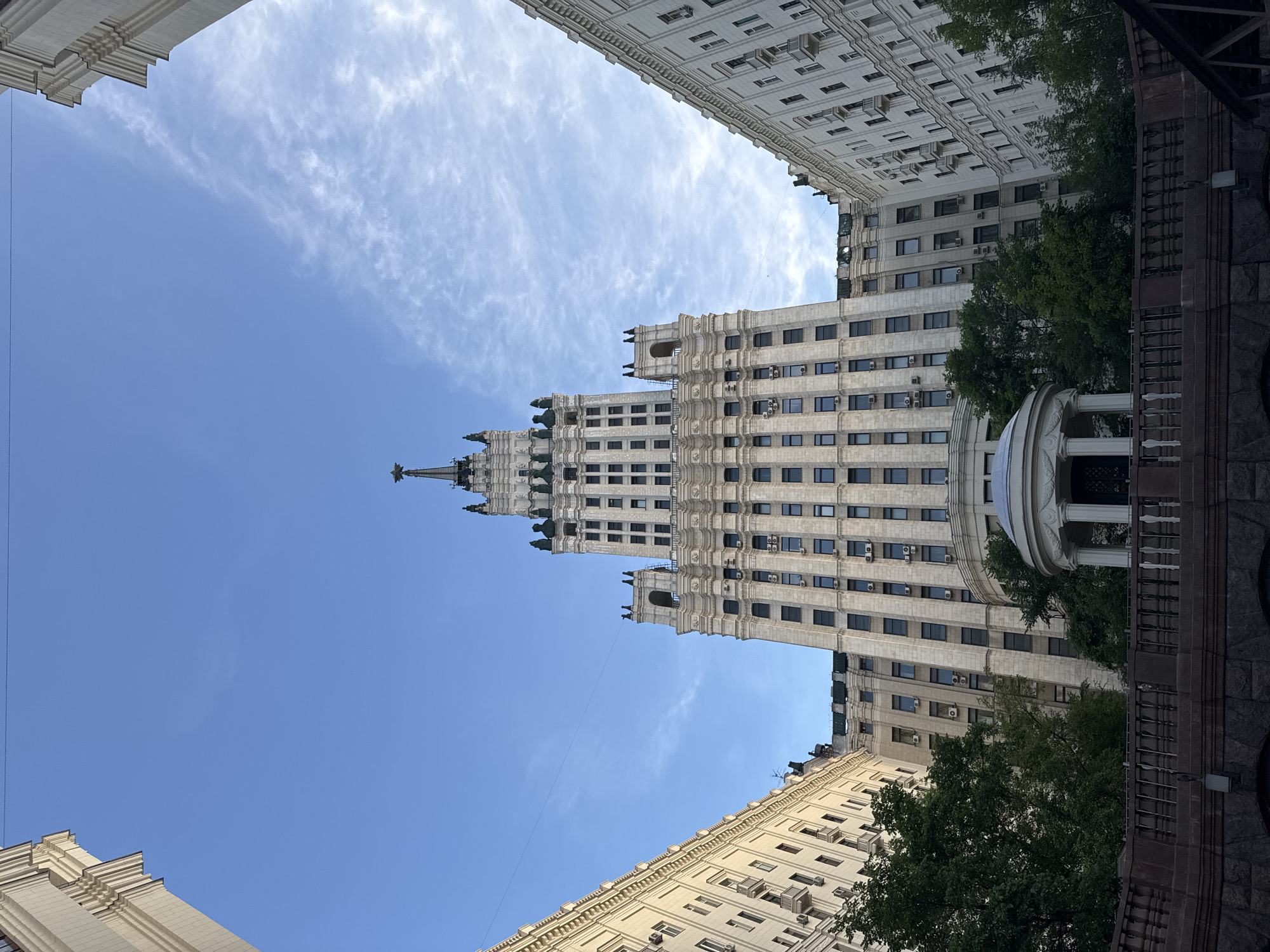 The image captures a low-angle view of the Kotelnicheskaya Embankment Building, a prominent Stalinist skyscraper located in Moscow, Russia. The building, constructed from light-colored stone, dominates the frame, its ornate facade stretching upwards towards a bright blue sky dotted with wispy white clouds. The central spire, topped with a red star, is clearly visible. Flanking the main building are two lower, symmetrical wings that curve inwards, framing the central structure and creating a sense of grandeur. Trees with dark green foliage are visible at the base of the buildings, providing a contrast to the architectural elements. In the foreground, a dark stone embankment with a decorative railing can be seen. The overall scene suggests an urban environment with imposing architecture under a clear sky.