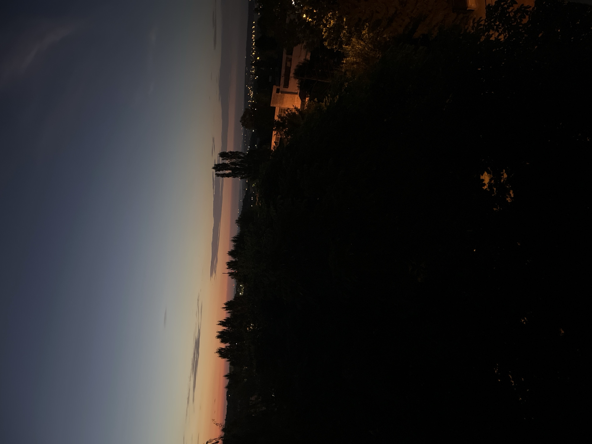 The image depicts a nighttime scene with a twilight sky. The sky transitions from a deep blue at the top to soft hues of orange and pink near the horizon, indicating it is either dusk or dawn. Silhouetted against the sky are trees and a building. The building appears to be a light-colored, multi-story structure, possibly a house or a small apartment complex, with lights visible in some of its windows. Scattered lights from a distant town or city are also visible along the horizon. The foreground is dominated by dark masses of trees, suggesting a natural or semi-natural setting like a park or a backyard overlooking a landscape. The overall mood is tranquil and serene, capturing the quiet beauty of the transition between day and night. There is no visible text in the image.