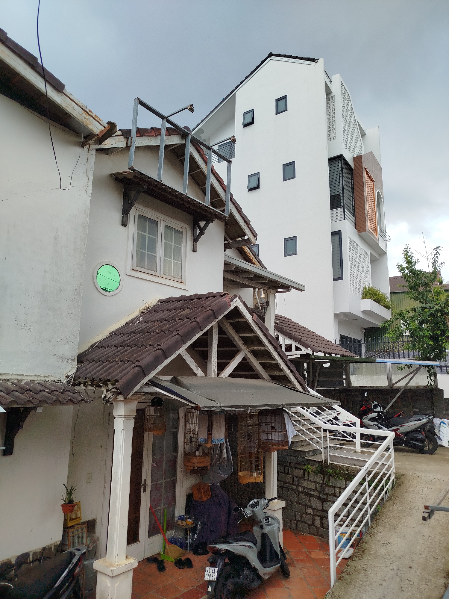This image presents a residential scene in Da Lat, Vietnam, under an overcast sky, showcasing a blend of traditional and modern architecture. In the foreground and to the left is an older, two-story white house with a distinctive brown tiled roof. Its facade exhibits some signs of age, including cracks and rusty guttering. A bright green circular window and a standard multi-pane window are visible on its upper floor. A metal frame structure, possibly for a balcony or an extension, protrudes from the upper level.
The ground floor of this house features a covered porch area where several traditional wooden bird cages, some containing birds, hang from the eaves. A sturdy white column supports the porch roof, and a white wooden door is visible behind a broom and slippers placed on the orange tiled ground. A gray Honda Vision scooter, bearing the Vietnamese license plate "49-AA 130.61," is parked prominently on these tiles. Further back and to the right, a white metal railing guides a stone-walled path or driveway, where other motorbikes, including a red and white model, are parked.
Dominating the background and rising behind the older structure is a taller, more modern white building. This contemporary design features small, dark square windows arranged irregularly, alongside decorative perforated sections on its facade, including a white latticework pattern and an arched section with orange-brown textured panels. A small tree with green foliage stands to the right of this modern building. The scene depicts everyday life in a lived-in neighborhood, characterized by parked vehicles, domestic bird-keeping, and a visual dialogue between different architectural eras. The cloudy conditions indicate daytime without direct sunlight.