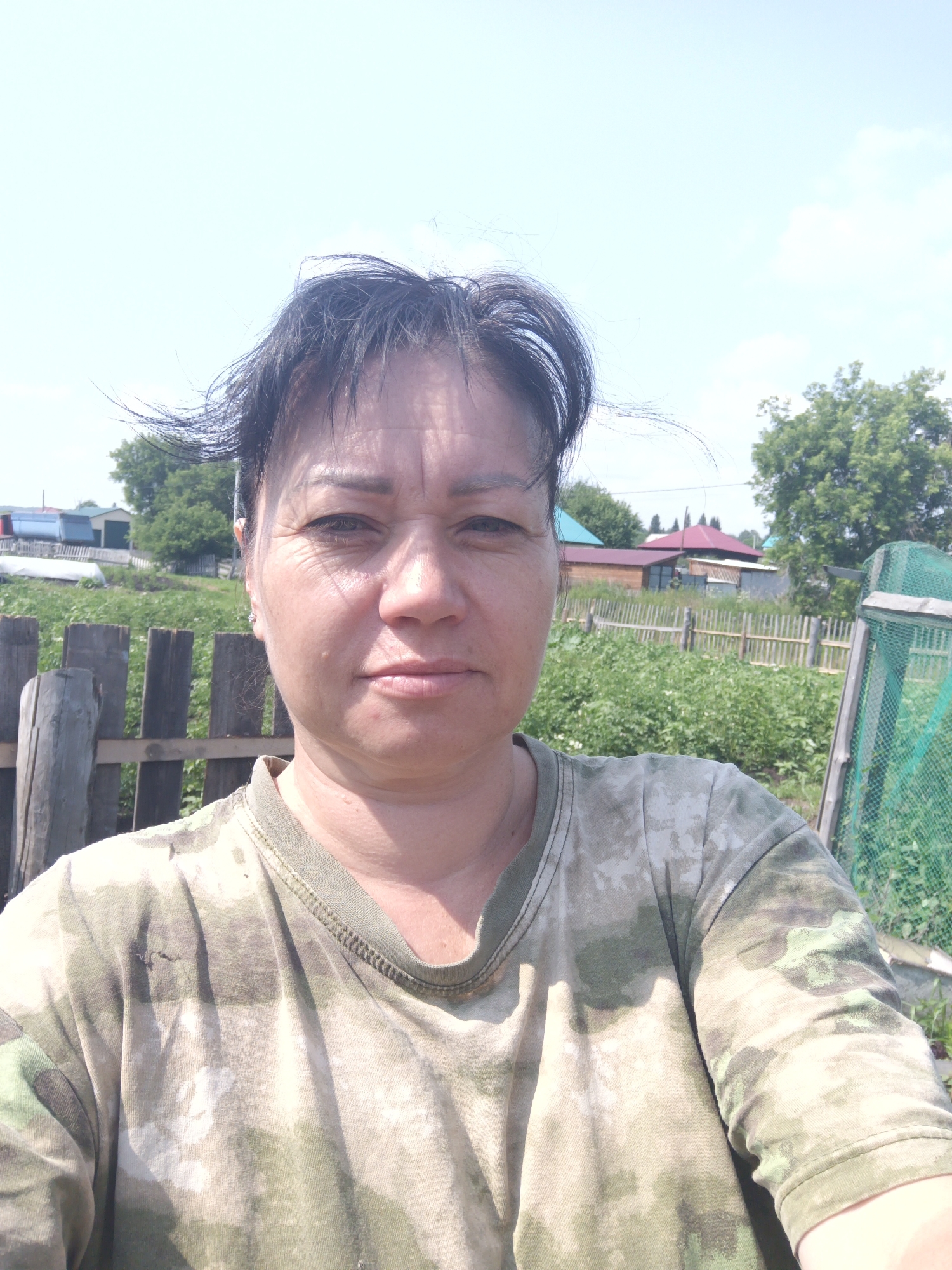 A middle-aged woman with dark hair, wearing a camouflage t-shirt, stands in what appears to be a rural setting. She is looking directly at the camera, with a slight smile. The background shows a wooden fence, green foliage, and several buildings with red and blue roofs, characteristic of a village or dacha settlement. The sky is clear and blue with a few wispy clouds. The scene suggests a warm, sunny day. Based on the architectural style and the general atmosphere, this could be the city of Zalesovo, Russia, a location known for its rural landscapes and traditional housing.