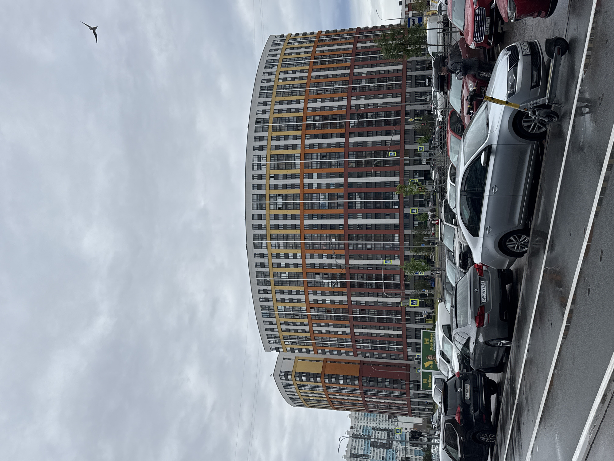 The image depicts a street scene with a large, modern apartment building featuring a colorful facade in St. Petersburg, Russia. The building is multi-storied, with alternating horizontal bands of white, yellow, orange, and red. In the foreground is a row of parked cars of various makes and models, including a silver Skoda, a gray BMW, and a black car with a license plate reading "E711HT 198," among others. There is also a silver electric scooter parked beside the cars. A street sign is visible behind the cars, and in the top left corner of the frame, a bird is flying in the overcast sky. The street is wet, suggesting recent rain.
