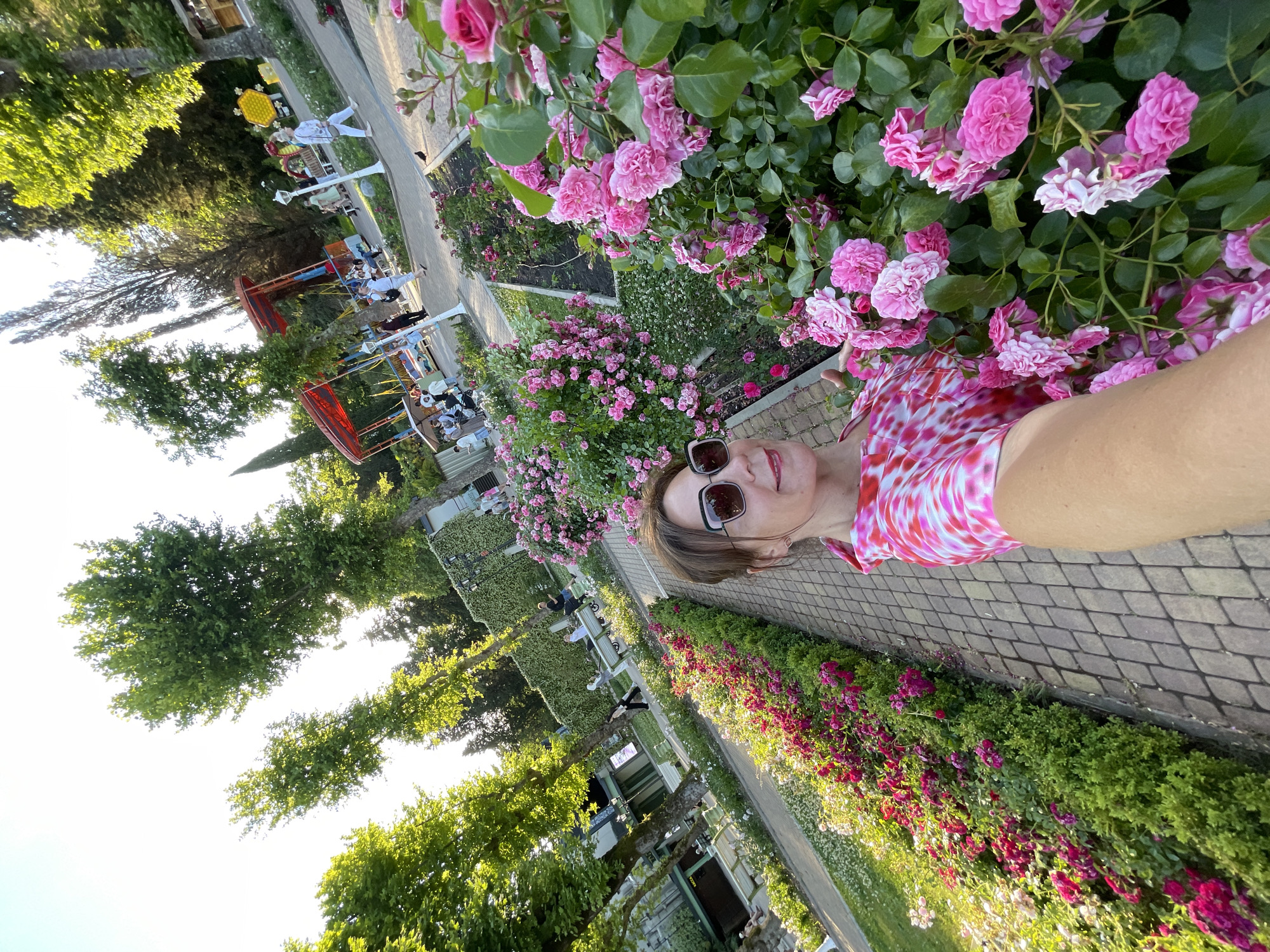 The image shows a woman taking a selfie in a rose garden located in Sochi, Russia. The woman, wearing a pink and white tie-dye top and sunglasses, is standing in front of a dense patch of pink roses. To her left, a paved walkway lines a garden bed filled with a variety of pink and red roses and greenery. In the background, several people are visible walking and sitting in a park-like setting with trees, benches, and children's amusement rides. The overall scene suggests a bright, sunny day in a public garden.
