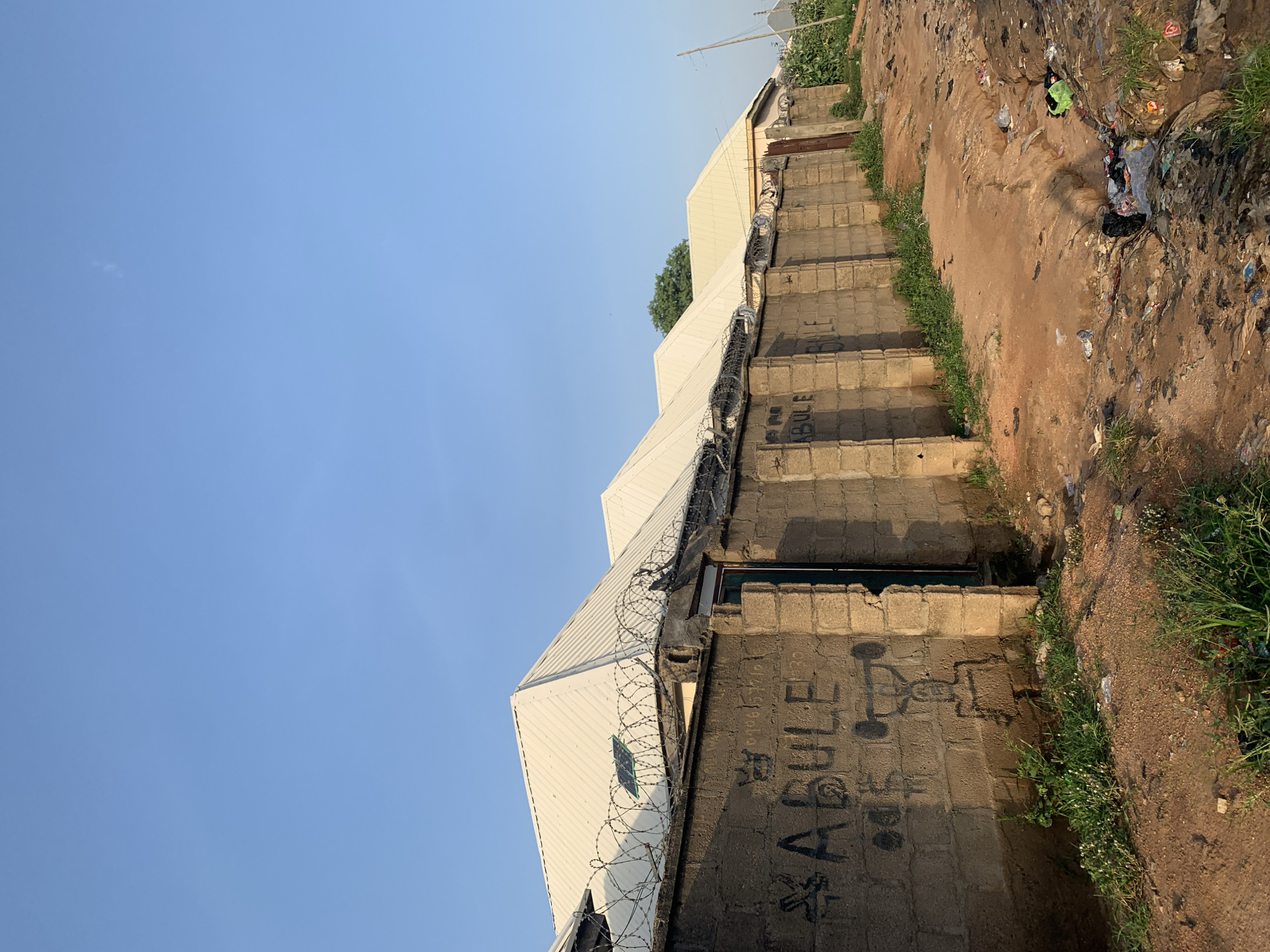 The image depicts a scene in Jauro Yinu, Nigeria, focusing on a residential area and its immediate surroundings. A long, weathered concrete block wall stretches across the frame, topped with barbed wire fencing, suggesting a measure of security. Graffiti, including the word "ABULE" and a crudely drawn figure holding weights, is visible on the wall. Behind the wall, several buildings with white, corrugated metal roofs are lined up, suggesting a row of houses or an estate. A small solar panel is mounted on one of the roofs. The ground in front of the wall is a dirt path with patches of grass. To the right of the wall and path, a significant amount of refuse and litter is scattered, indicating a lack of proper waste management in the area. The sky above is a clear, pale blue, indicating a daytime scene under good weather conditions. The overall impression is one of a developing urban or semi-urban environment with a contrast between residential structures and the presence of waste.