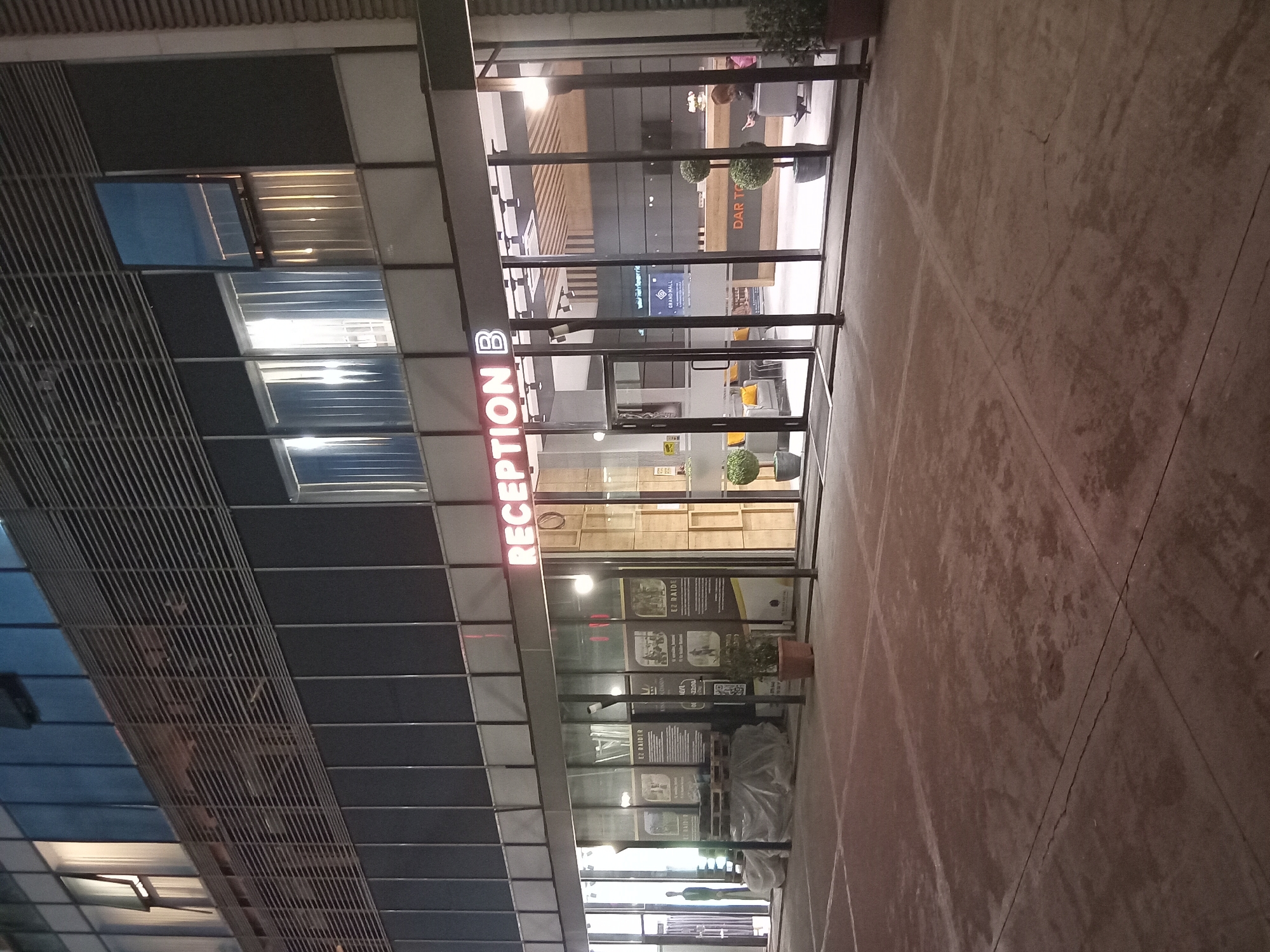 The image shows the exterior of a building, likely located in Batumi, Georgia, based on the presence of a sign for "Grand Mall" (potentially referencing the Batumi Grand Mall). The building has a modern architectural style with a facade featuring a grid-like pattern and large glass windows/doors. A prominent sign above the entrance reads "RECEPTION B" in red neon letters. Inside, there are glimpses of a retail space or lobby, with furniture and decorations visible. Several posters or advertisements with "EZ RAIDER" printed on them are visible. Outside, the ground appears to be paved. There is what appears to be wrapped goods. No individuals are clearly identifiable in the immediate foreground of the entrance.