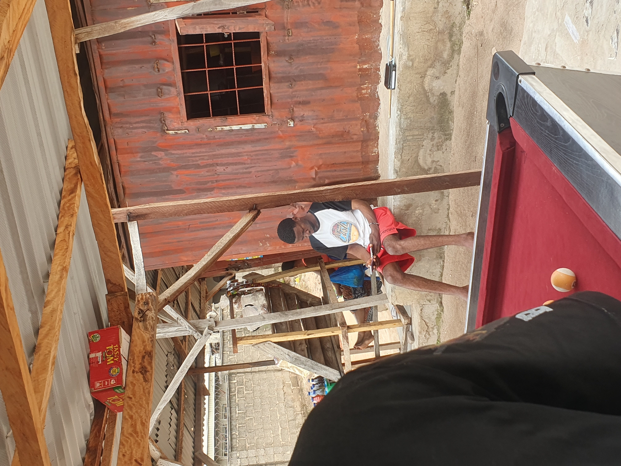 The image depicts an outdoor scene, possibly a recreational area or a makeshift establishment, in Lokoja, Nigeria. In the foreground, a portion of a red billiards table is visible. Behind the table, a young Black man is seated on a concrete ledge, looking down at a smartphone he is holding. He is wearing a white t-shirt with black sleeves and red shorts. To his left, a corrugated metal building with a barred window stands. Above the man and building, a wooden roof structure with exposed beams and a corrugated metal roof is apparent. In the background, a brick wall and some stairs can be seen. A red box with "TASTY TOM" branding is placed on an overhead wooden beam. The ground appears to be sandy or dirt. The overall impression is of a casual, somewhat rustic environment.