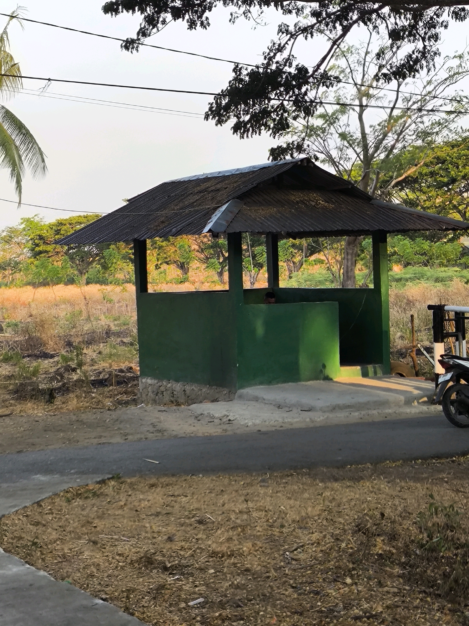 In an outdoor setting in Unknown, Indonesia, a small, open-sided, green structure with a corrugated metal roof stands on a concrete base. The structure appears to be a simple shelter or guard post. A person is visible inside the shelter, partially obscured by the walls. The surrounding environment is dry and consists of sparse vegetation, including tall grasses and some trees in the background. Electrical wires are visible overhead, strung between poles or trees. A road or paved pathway runs in front of the shelter, and a motorcycle is parked to the right of the structure, suggesting it is accessible by vehicle. The scene is lit by natural sunlight, casting shadows on the ground.