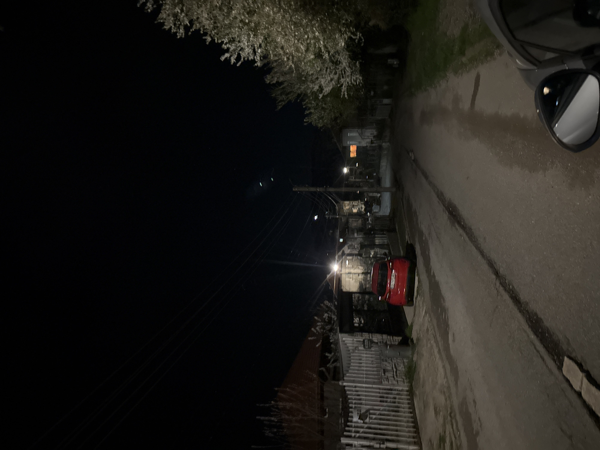 At night in Berkovitsa, Bulgaria, a street scene shows a narrow road alongside a building with a fence. A red car is parked next to the fence. Utility poles with overhead wires traverse the dark sky. The street is illuminated by a streetlight. A tree branch extends into the upper left of the frame. A car's side mirror is visible in the upper right corner.