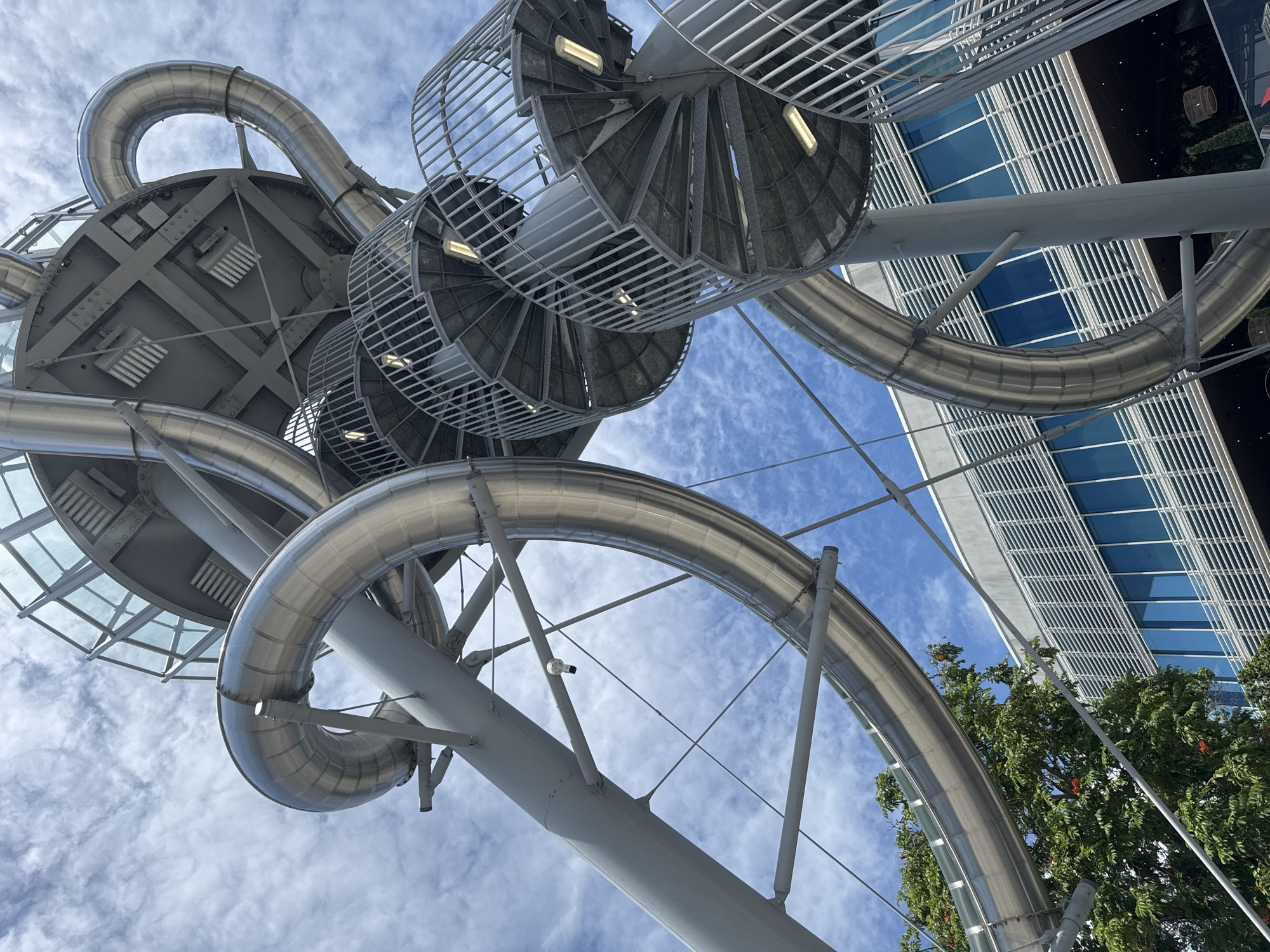 This is a low-angle, close-up shot of a large, metallic amusement park slide structure against a cloudy blue sky. The slide is a winding, tubular structure made of polished metal, supported by a complex network of gray metal beams and columns. Several spiral staircases with metal railings lead up to the platforms of the slide. The overall impression is one of intricate engineering and a playful, adventurous atmosphere. In the background, a modern building with a glass facade and horizontal louvers is visible, partially obscured by the slide structure and a lush green tree. The image appears to be taken in Aventura, Florida, United States of America.