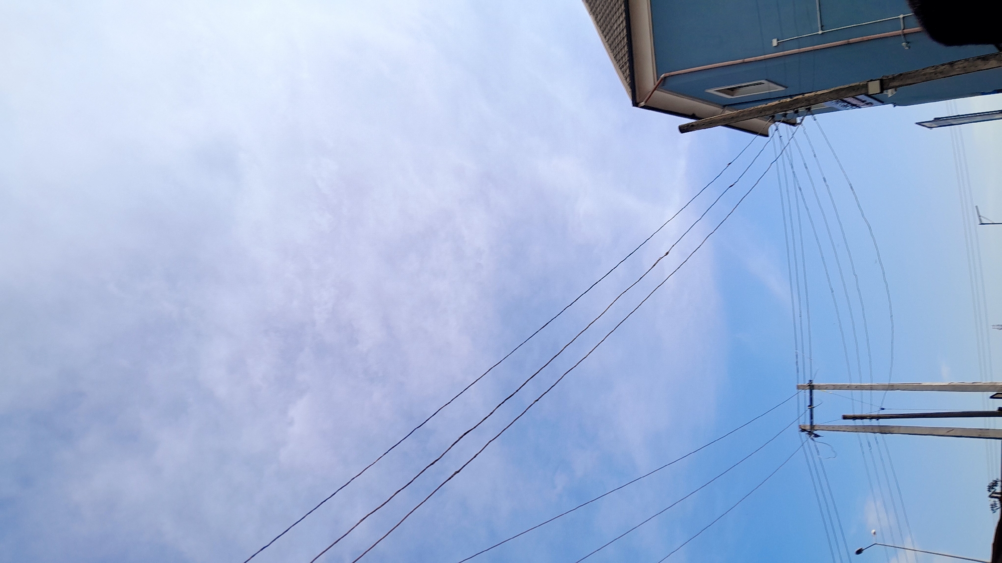 The image shows a view looking upwards toward the sky in Elelenwo, Nigeria. Dominating the upper right corner of the frame is part of a blue building. Numerous power lines stretch across the sky, connecting to utility poles. The sky is a mix of blue and white, partially obscured by clouds.