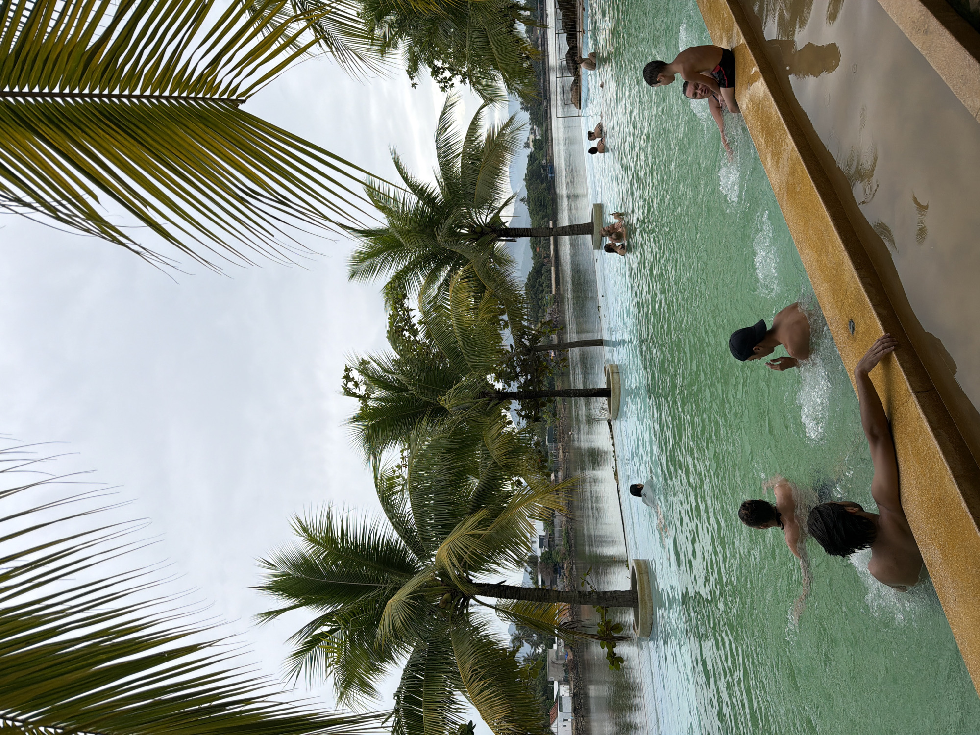 The image depicts a serene swimming pool area with a scenic river backdrop, likely in Nha Trang, Vietnam. Palm trees line the pool, their fronds creating a natural canopy against a cloudy sky. Several people are visible in the greenish-blue water, some swimming, others lounging and interacting. In the foreground, a person with dark hair rests their arm on the pool's edge, their body partially submerged. To their right, another person is seated on the edge of the pool, looking towards the viewer. Further in the pool, individuals are scattered, enjoying the water. The river extends into the background, with a lush green landscape and distant hills under the overcast sky. Buildings and structures are visible along the riverbank, suggesting proximity to a town or village. The overall atmosphere is one of relaxation and leisure in a tropical environment.