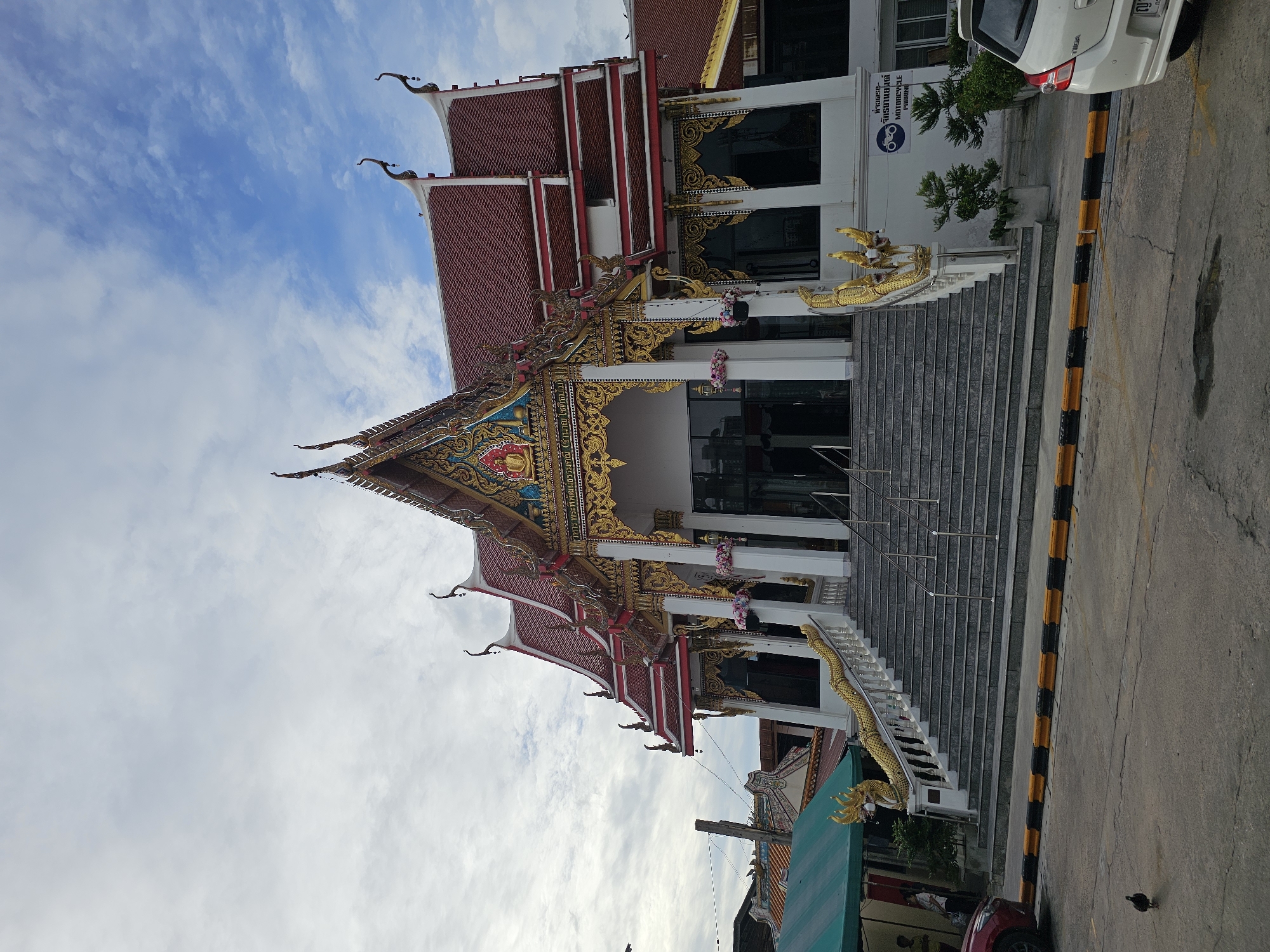 The image shows a temple in Bangkok, Thailand, with a grand staircase leading up to the entrance. The temple features ornate golden decorations and a red tiled roof, typical of Thai Buddhist architecture. A golden dragon sculpture flanks the staircase. In the foreground, a yellow and black striped barrier separates the temple grounds from a paved road where a red car and a white car are parked. A single bird is visible on the road. The sky above is partly cloudy with patches of blue. The overall scene depicts a vibrant and culturally significant location within the bustling city of Bangkok.